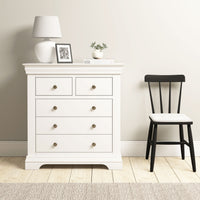 The Chalbury 2 Over 3 Chest of Drawers in Warm White is topped with decor and placed beside a black chair on a wooden floor against a light-colored wall.