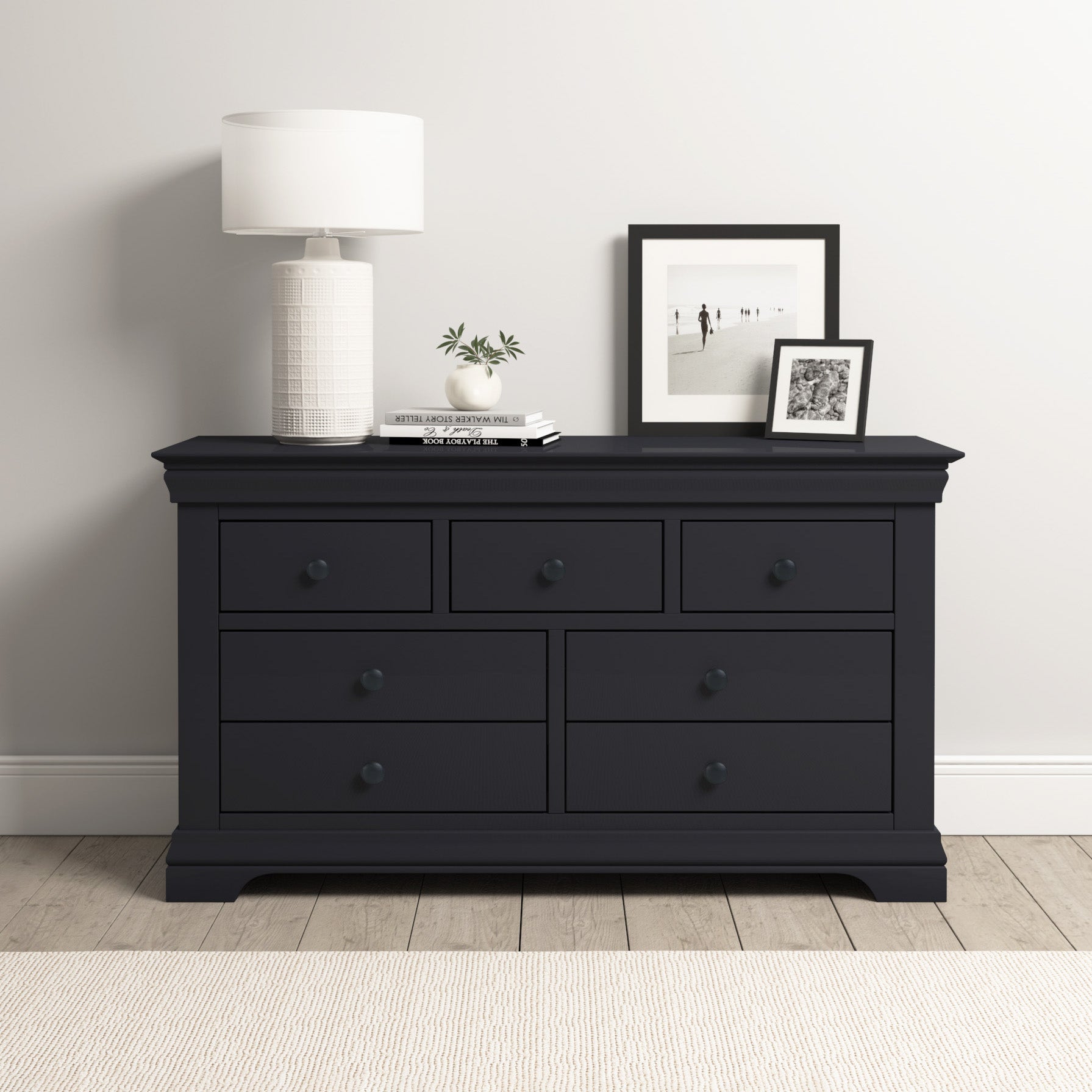 The Chalbury 3 Over 4 Chest of Drawers in Dusky Black offers modern storage with seven spacious drawers, shown topped with a white lamp, two framed photos, and a small plant.