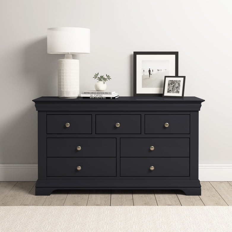 The Chalbury 3 Over 4 Chest of Drawers in Dusky Black has a white lamp, two photo frames, and a small plant on top, set against a pristine white wall.