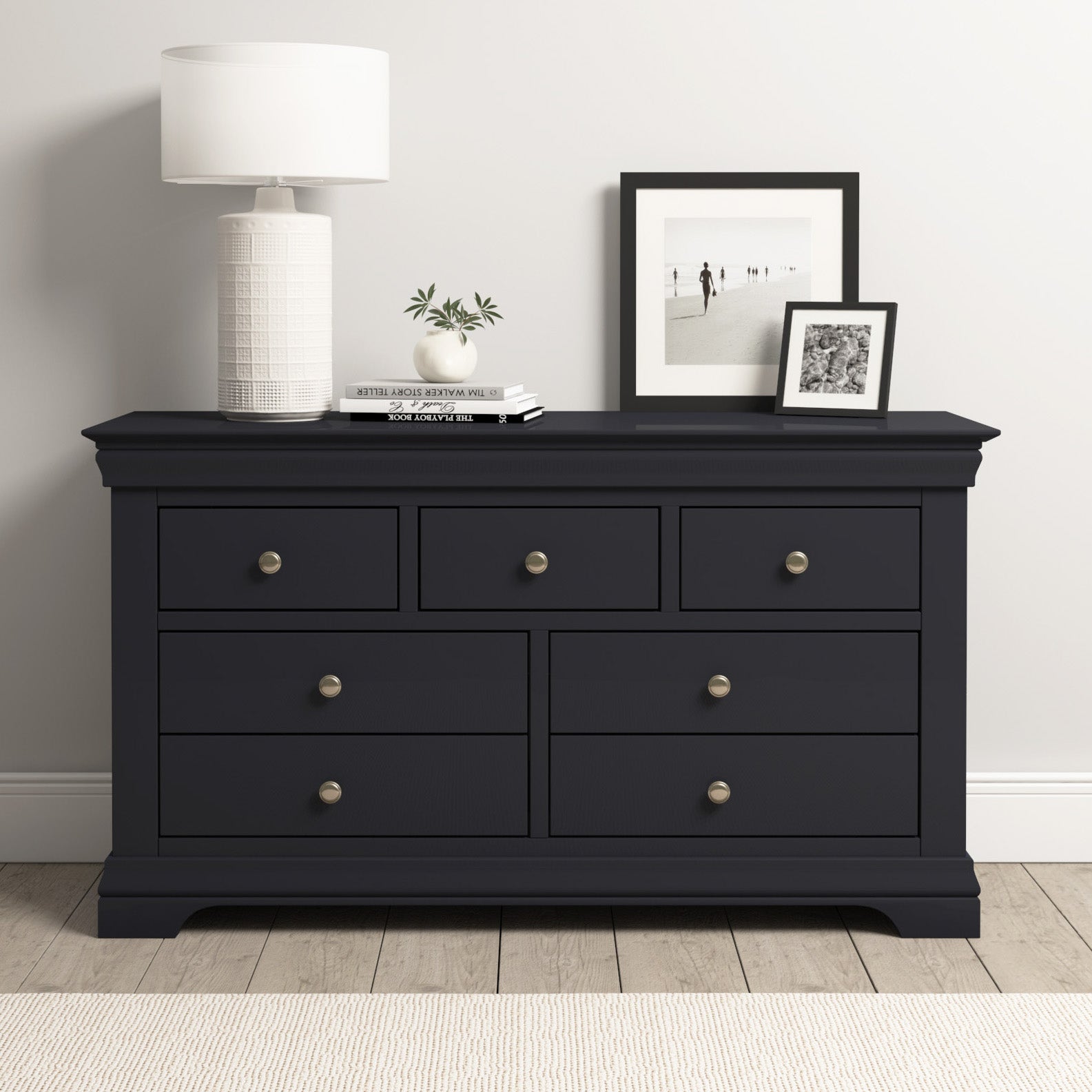 The Chalbury 3 Over 4 Chest of Drawers in dusky black features six roomy compartments and is styled with a lamp, books, plant, and framed photos on top—modern storage set against a crisp white wall.