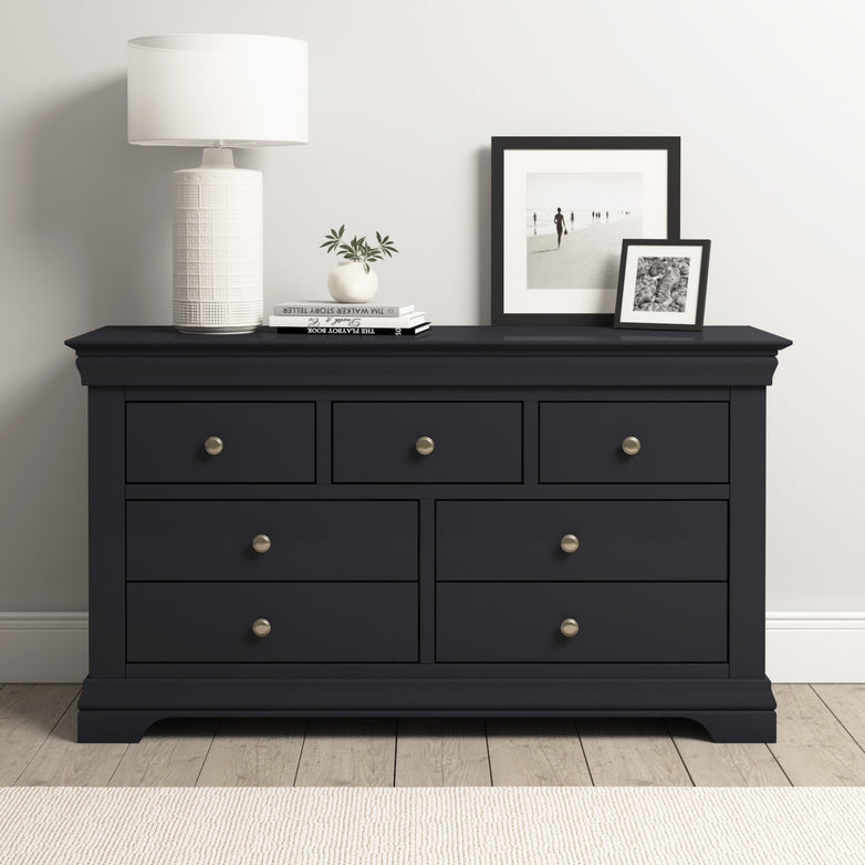 The Chalbury 3 Over 4 Chest of Drawers in dusky black features six roomy compartments and is styled with a lamp, books, plant, and framed photos on top—modern storage set against a crisp white wall.