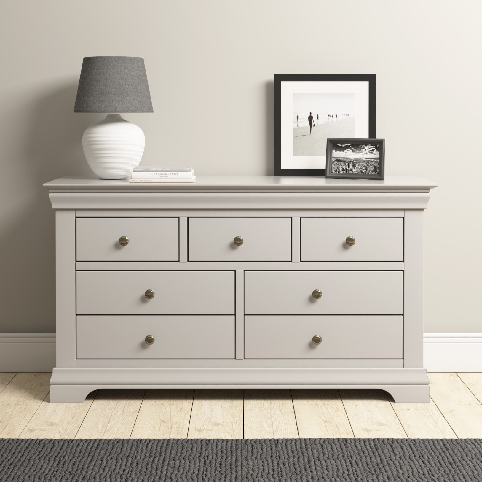 A Chalbury 3 Over 4 Chest of Drawers in Pebble Grey stands elegantly against a beige wall, adorned with a lamp and framed photos. Its sleek design provides functional storage on the warm wooden floor.