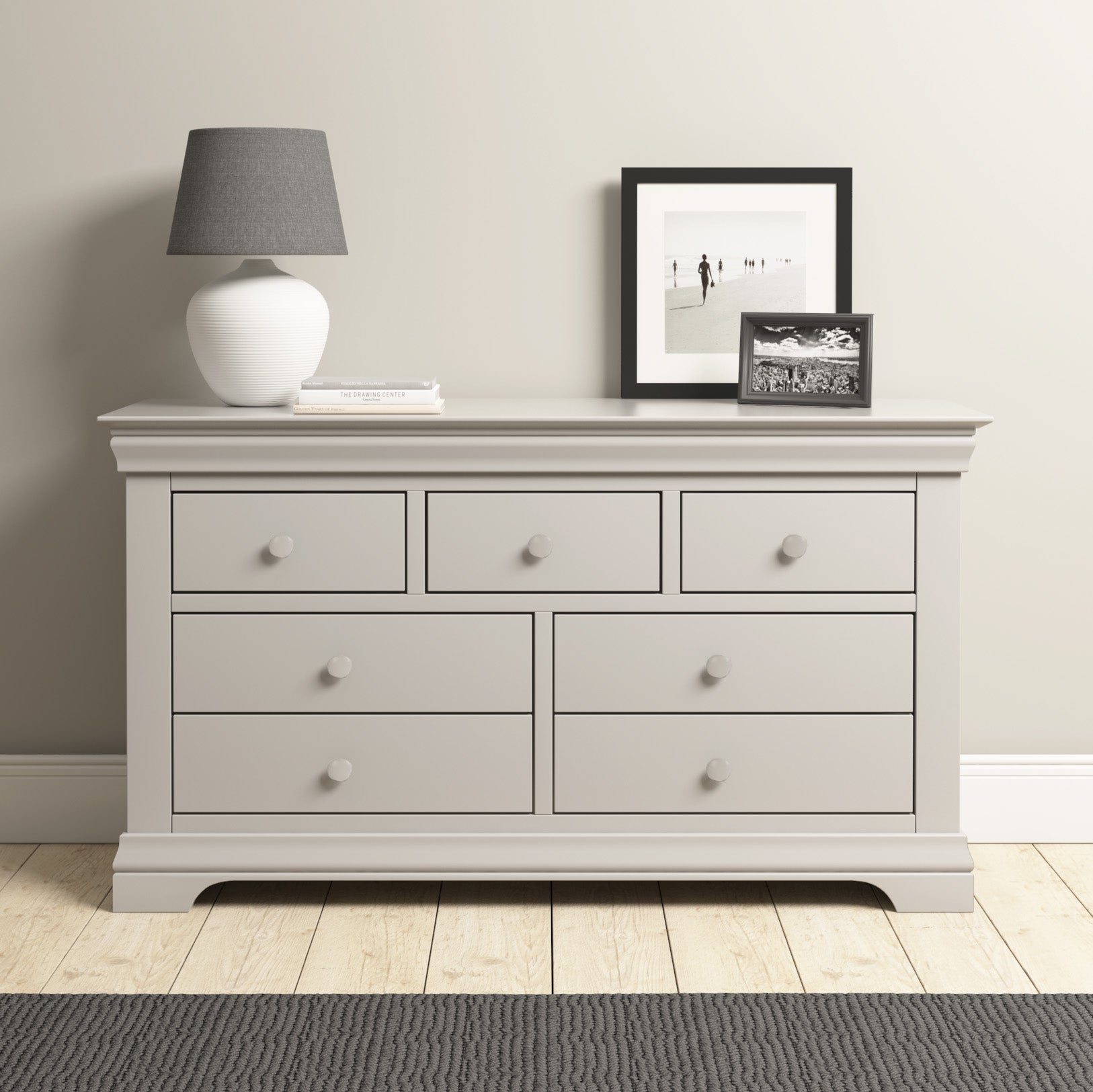 The Chalbury 3 Over 4 Chest of Drawers in pebble grey offers ample storage with seven drawers, topped by a white vase lamp, two framed photos, and a book. Set on a light wooden floor, it combines style and functionality beautifully.