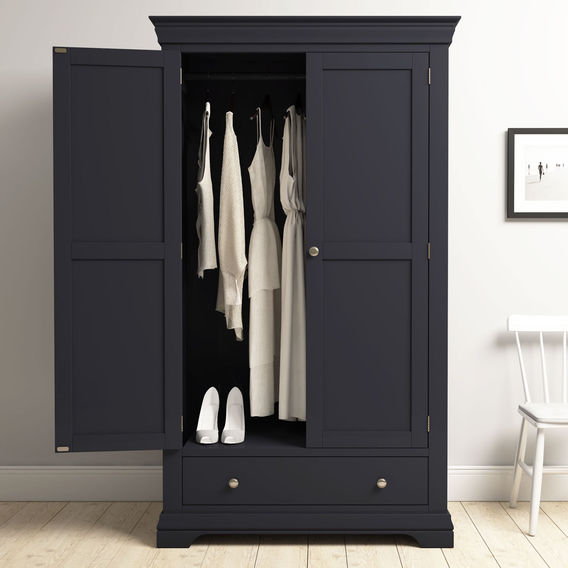 The Chalbury Double Wardrobe in Dusky Black stands open to display white dresses and shoes inside, providing elegant bedroom storage beside a chair and a photo on the wall.