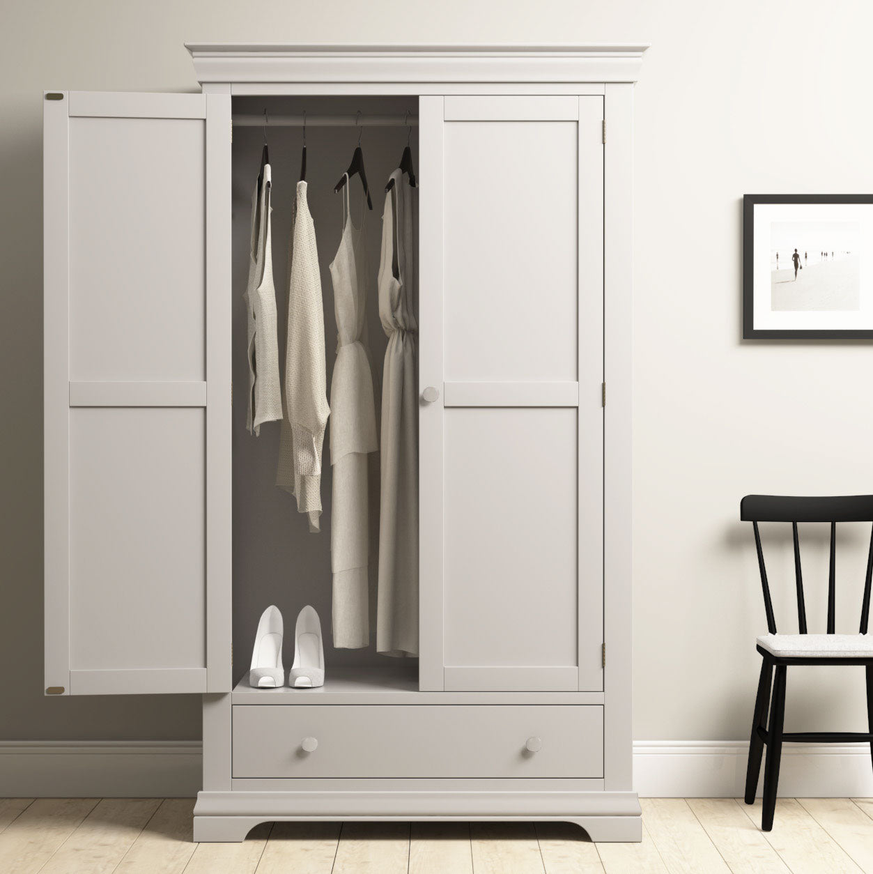 The Chalbury Double Wardrobe in Pebble Grey stands open, displaying hanging clothes, white shoes, and a drawer for elegant bedroom storage, next to a black chair.