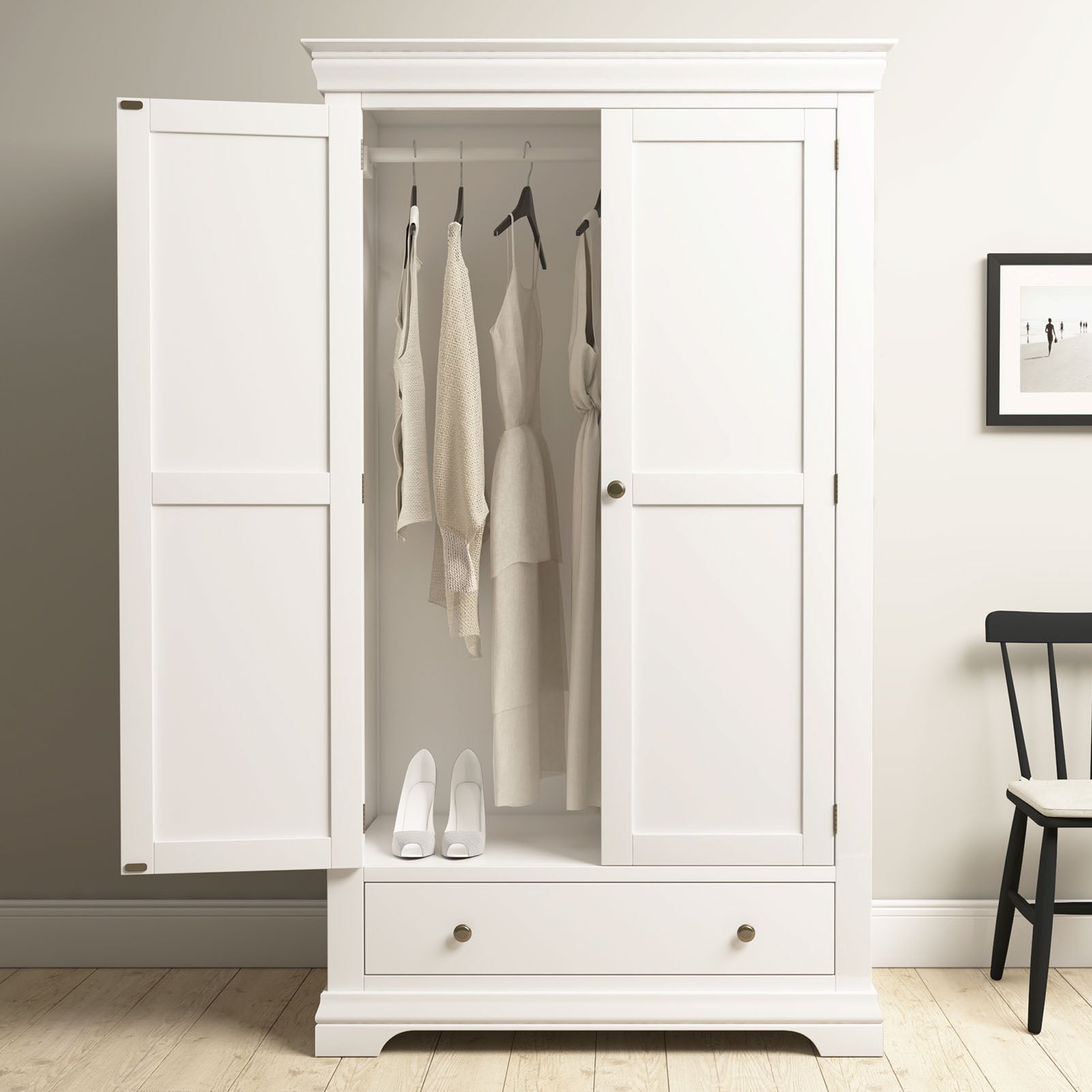 The Chalbury Double Wardrobe in Warm White provides generous bedroom storage with open doors, hanging beige clothes, white shoes underneath, and a bottom drawer. Its warm white finish suits any décor.