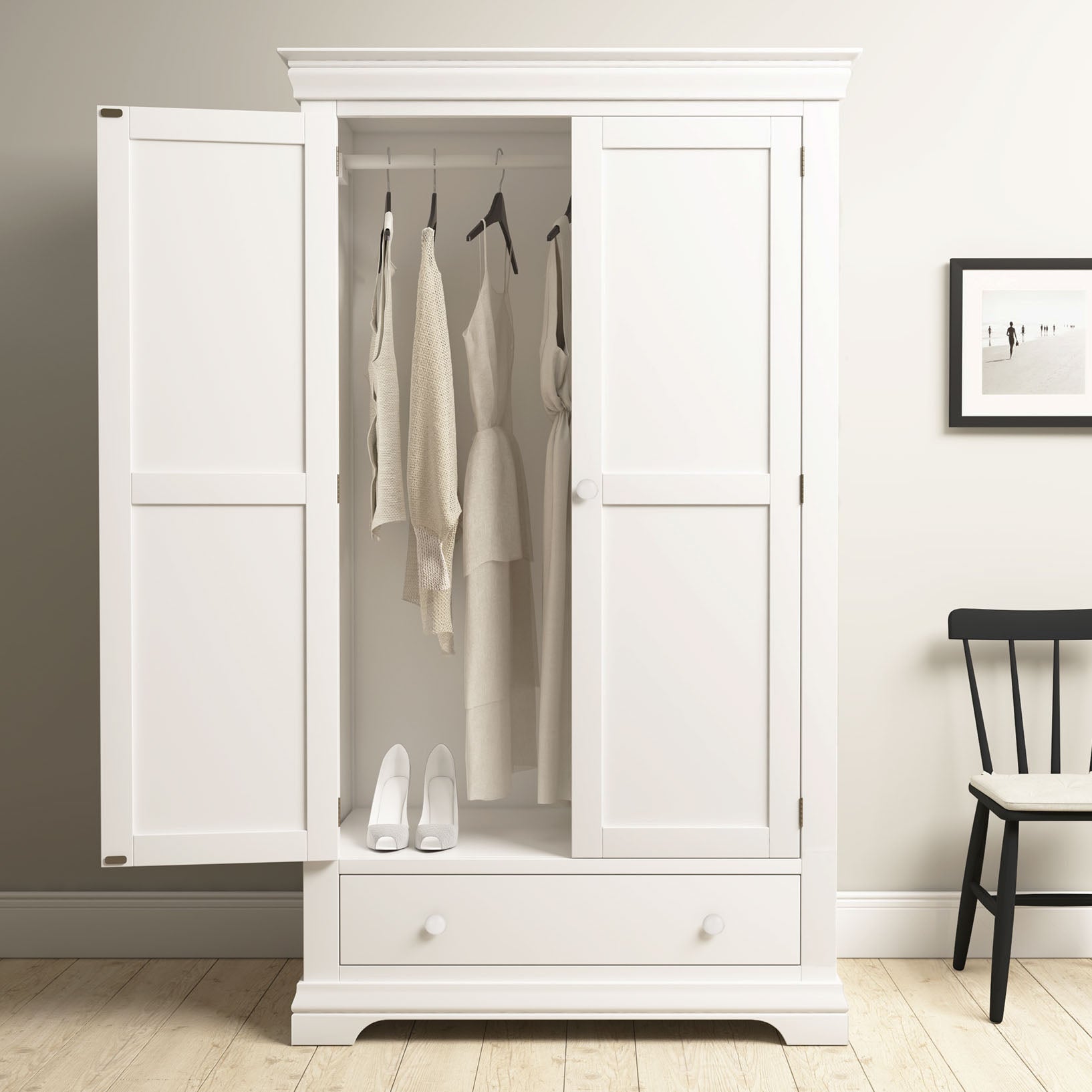 The Chalbury Double Wardrobe in Warm White features open doors with neutral clothes on hangers and white shoes inside, providing stylish bedroom storage. A chair and framed photo are placed nearby for a cozy touch.