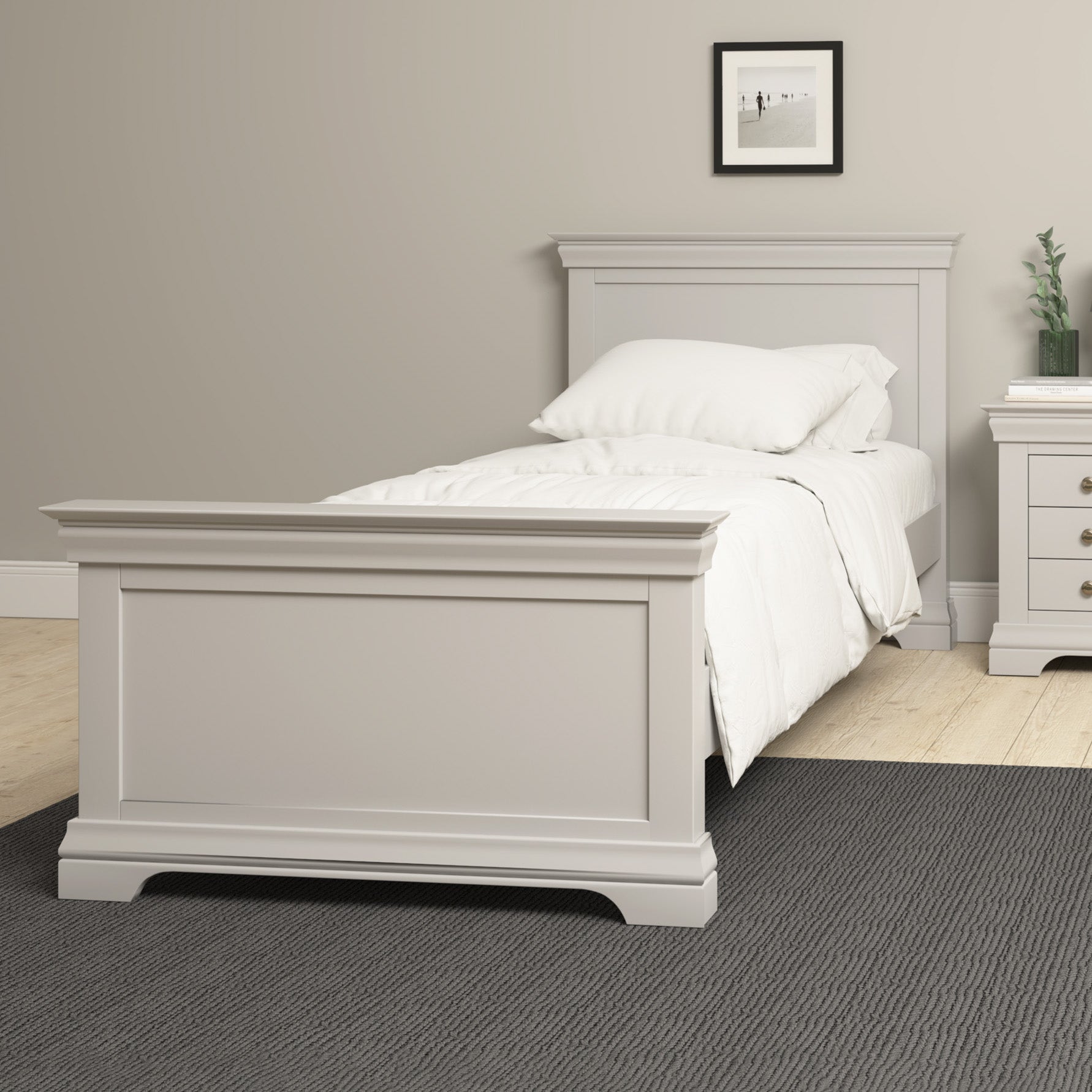 A neatly made bed with white bedding rests on a Chalbury 3ft High-End Single Bed Frame in Pebble Grey, accompanied by a nightstand, atop a Pebble Grey rug in a beige room exuding luxury design.
