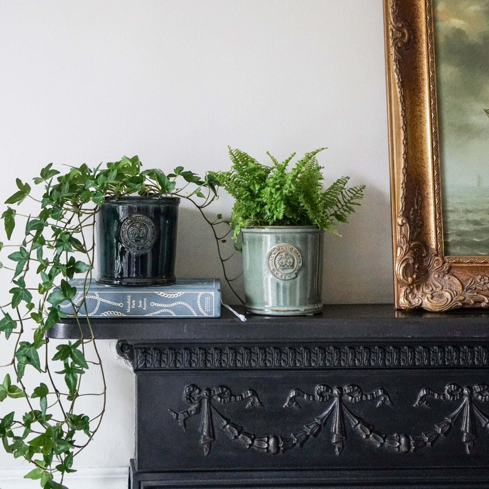 Two Ivyline Kew Reactive Glaze Emerald Planters sit on a black mantelpiece next to a framed painting, with one perched atop a book, embodying the elegant artistry of Kew Gardens.