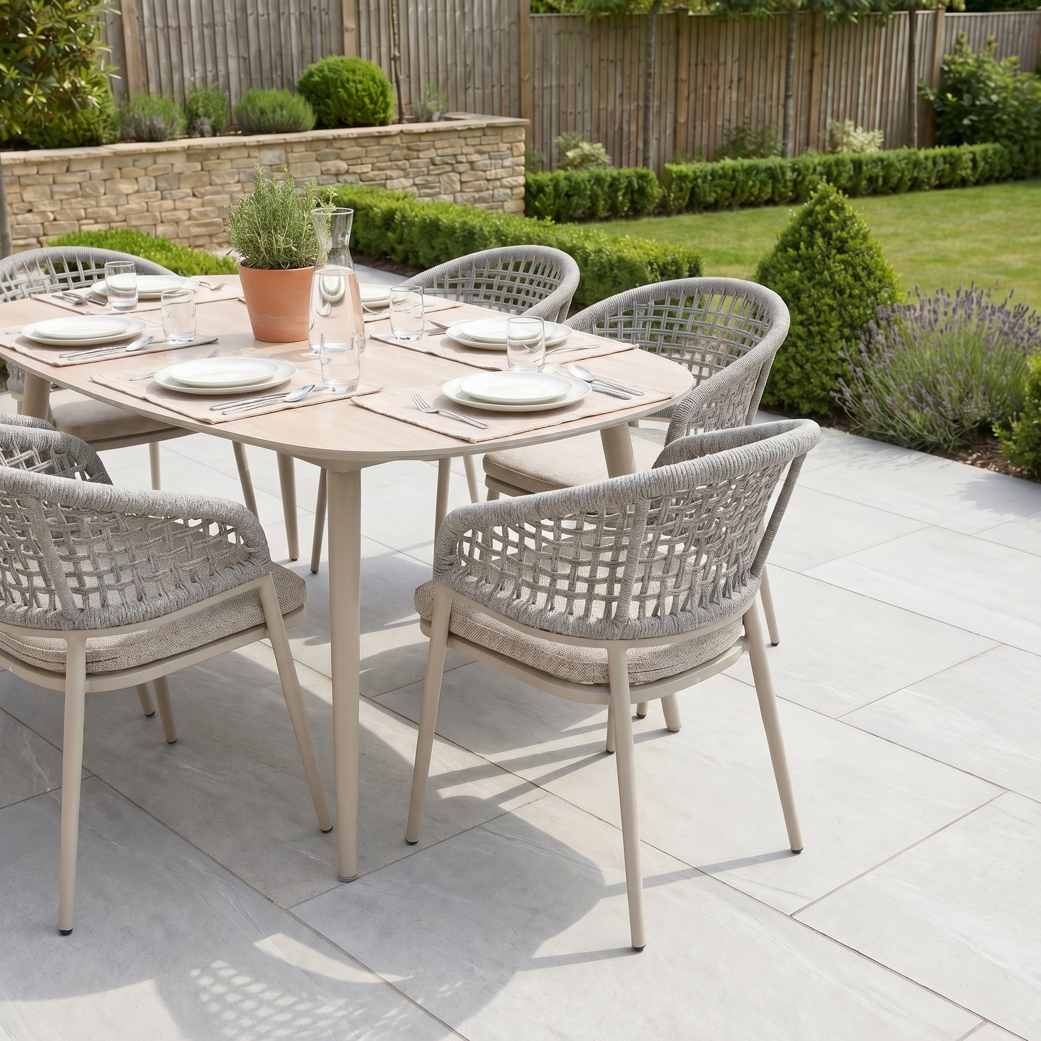 The Kalama 6 Seat Oval Rope Dining Set in Oatmeal brings comfort and style to your patio, creating the ideal outdoor dining arrangement amidst a well-kept garden with lush greenery and flowers.