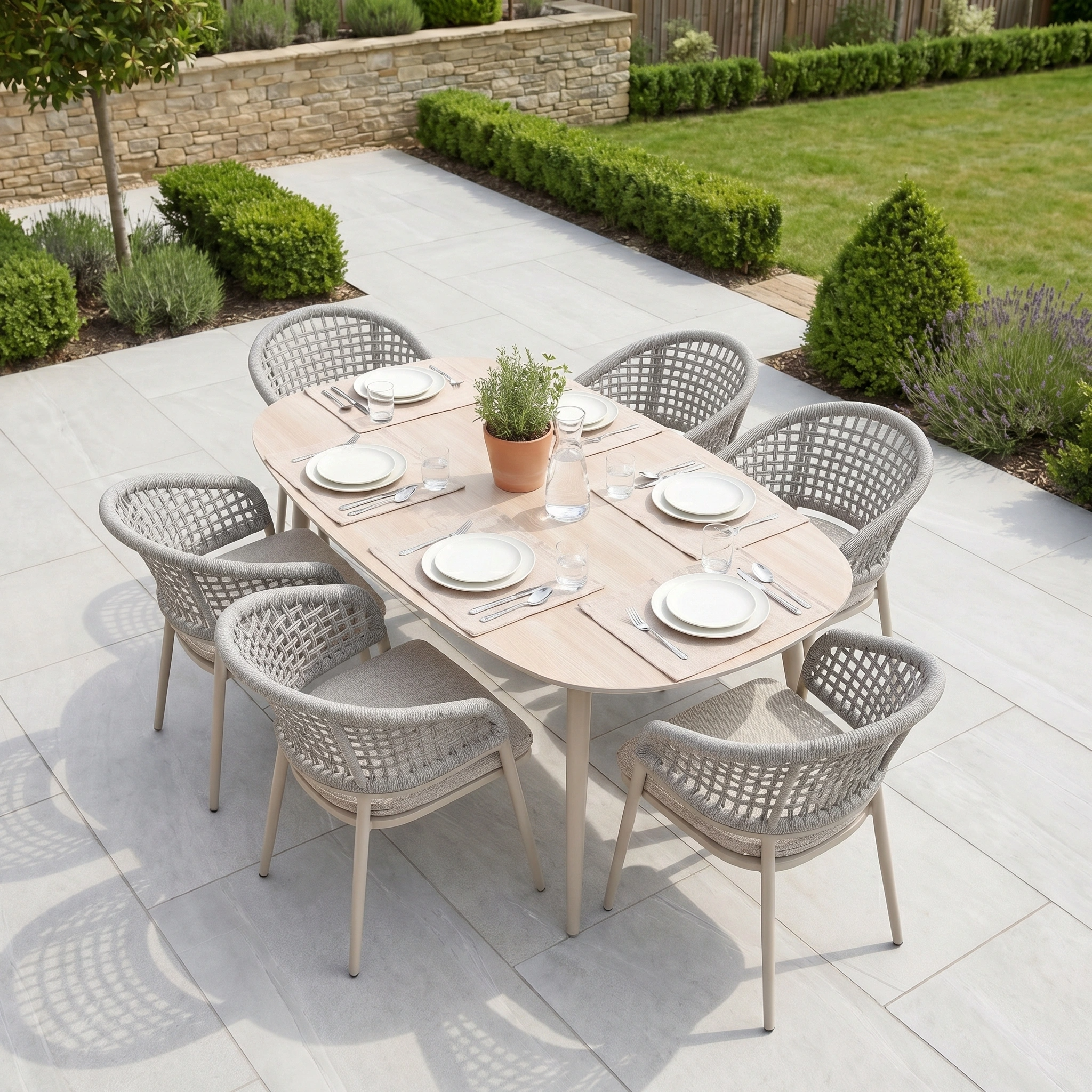 The Kalama 6 Seat Oval Rope Dining Set in Oatmeal, set with white plates and glassware, is surrounded by woven chairs and lush garden greenery—a perfect choice for stylish outdoor dining.