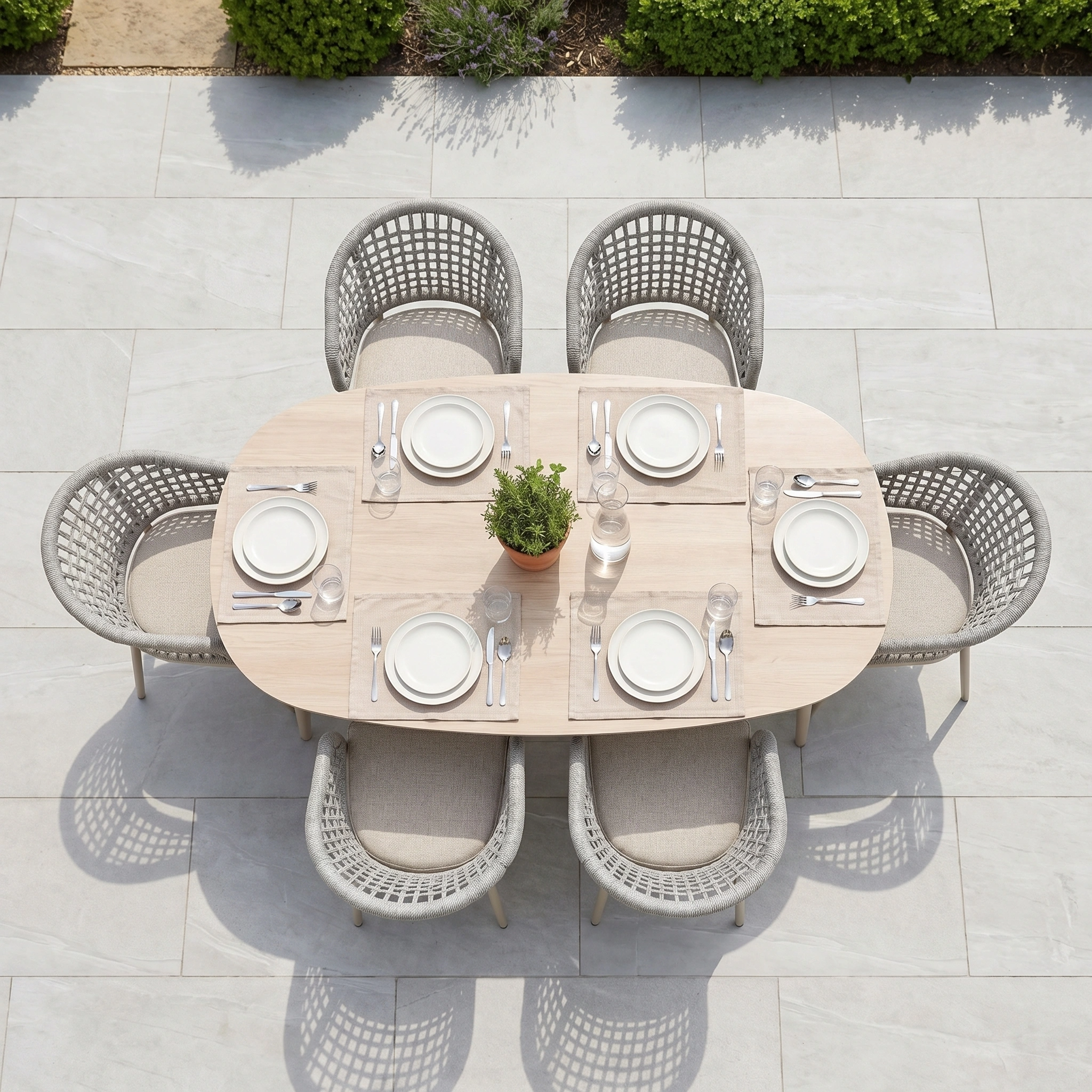 The Kalama 6 Seat Oval Rope Dining Set in Oatmeal features elegant garden furniture with seating for six, perfect for al fresco gatherings. Includes white plates, cutlery, glasses, and a potted plant centerpiece.