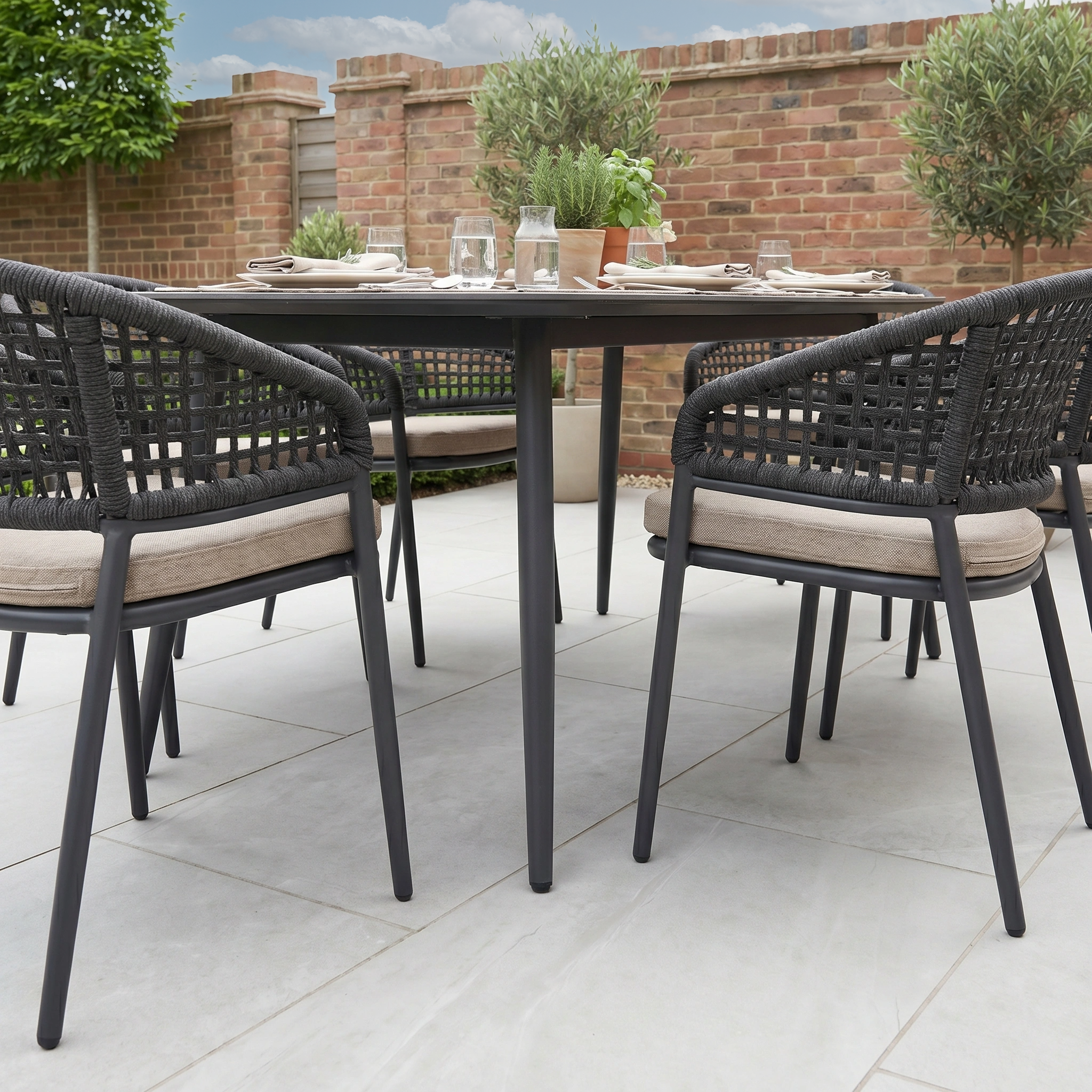 The Kalama 6 Seat Oval Rope Dining Set in Truffle, arranged on a patio with plates and glasses and surrounded by greenery, is perfect garden dining furniture for relaxed al fresco meals.