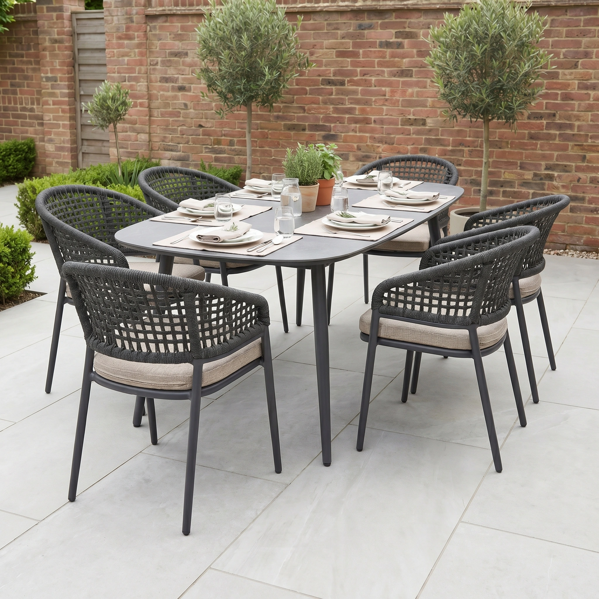 The Kalama 6 Seat Oval Rope Dining Set in Truffle features six cushioned chairs, ideal as stylish garden dining furniture for your patio surrounded by brick walls and potted plants.