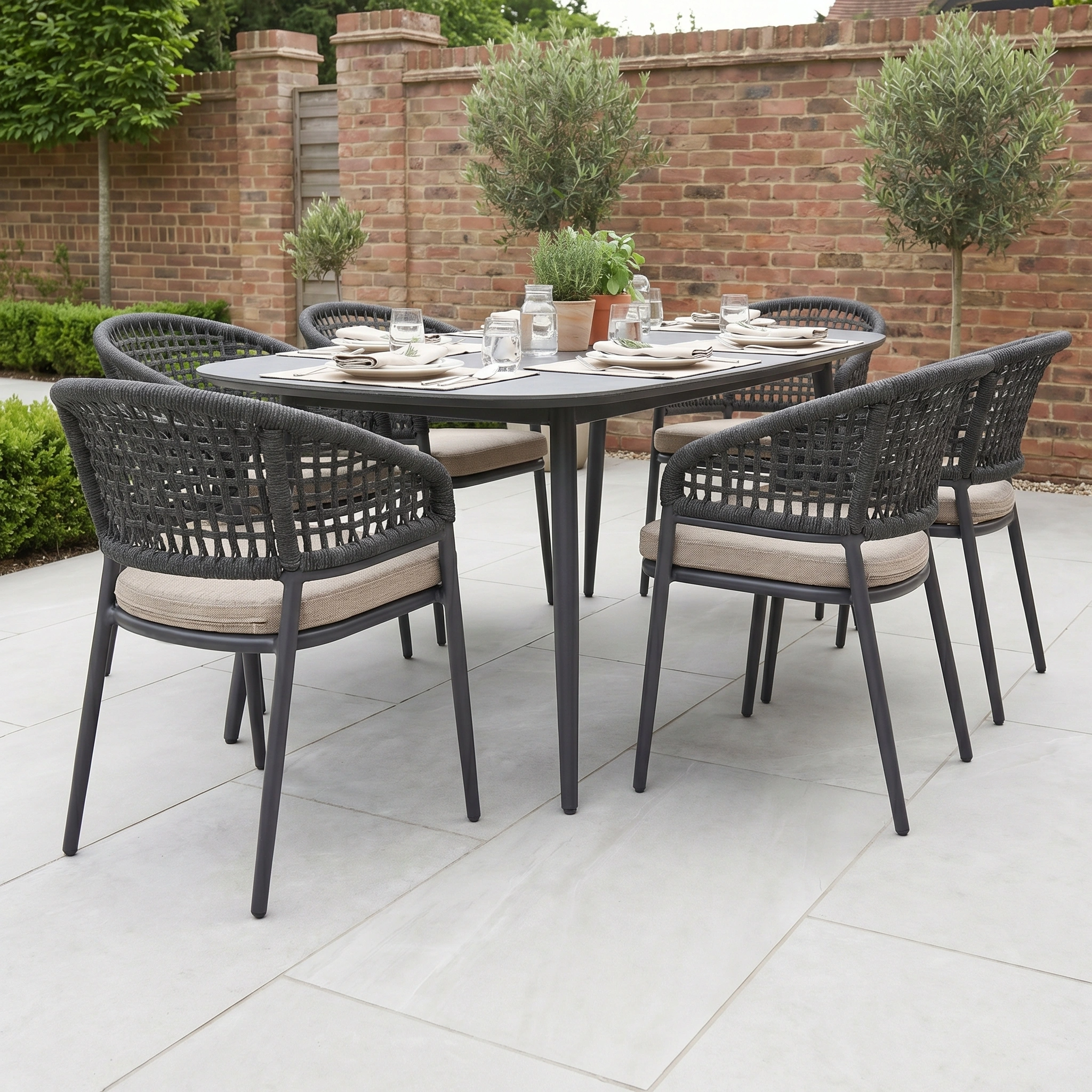 The Kalama 6 Seat Oval Rope Dining Set in Truffle features six woven chairs and a sleek oval table—ideal for stylish outdoor dining on your patio, surrounded by lush greenery.
