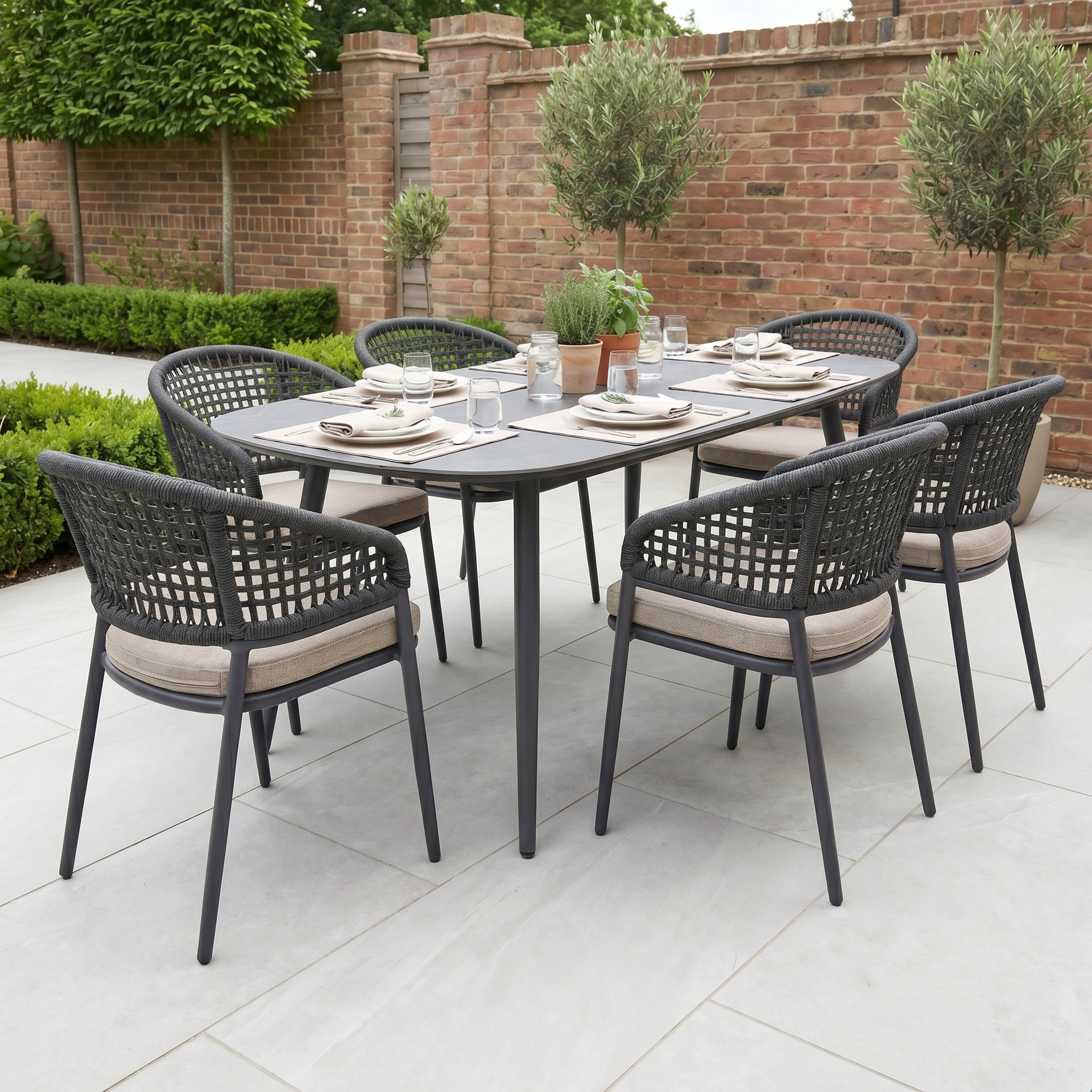 The Kalama 6 Seat Oval Rope Dining Set in Truffle features six chairs, perfect for outdoor dining in a lush garden surrounded by brick walls and greenery.