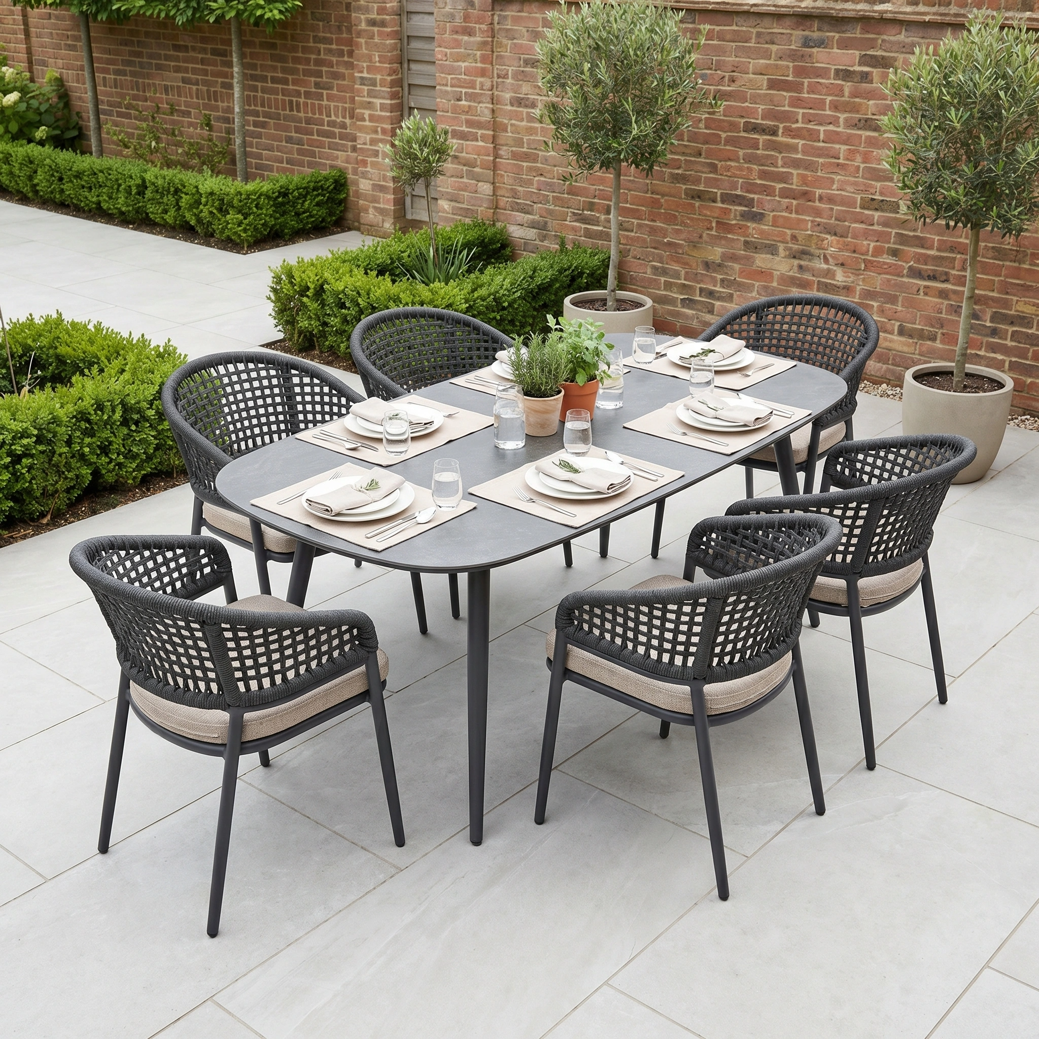 The Kalama 6 Seat Oval Rope Dining Set in Truffle, with black chairs, plates, glasses, and potted plants, creates a stylish patio atmosphere—ideal garden dining furniture for gatherings with family and friends.