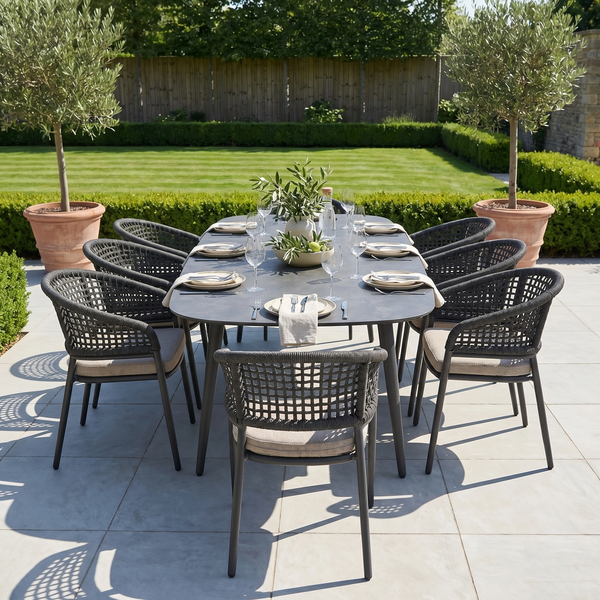 Outdoor dining set for eight on a patio, featuring the Kalama 8 Seat Oval Rope Dining Set in Truffle, made from sleek aluminium and rope, surrounded by potted trees and a lush green lawn.