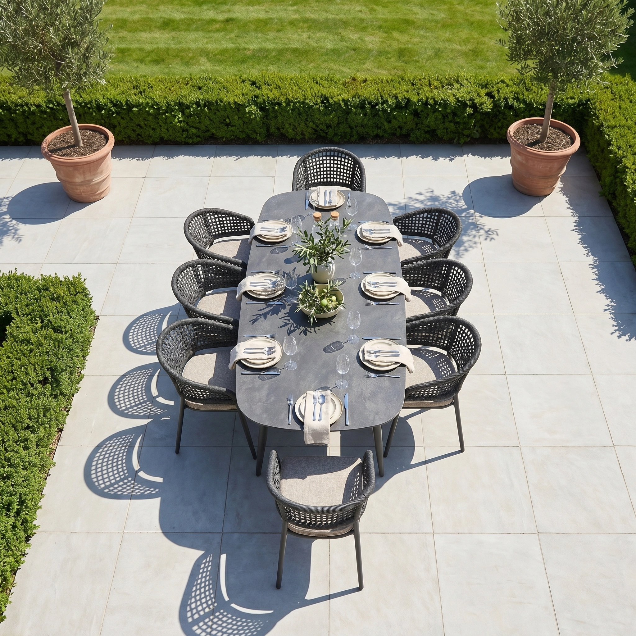 The Kalama 8 Seat Oval Rope Dining Set in Truffle offers stylish outdoor dining, ideal for enjoying meals on a tiled patio amid lush greenery.