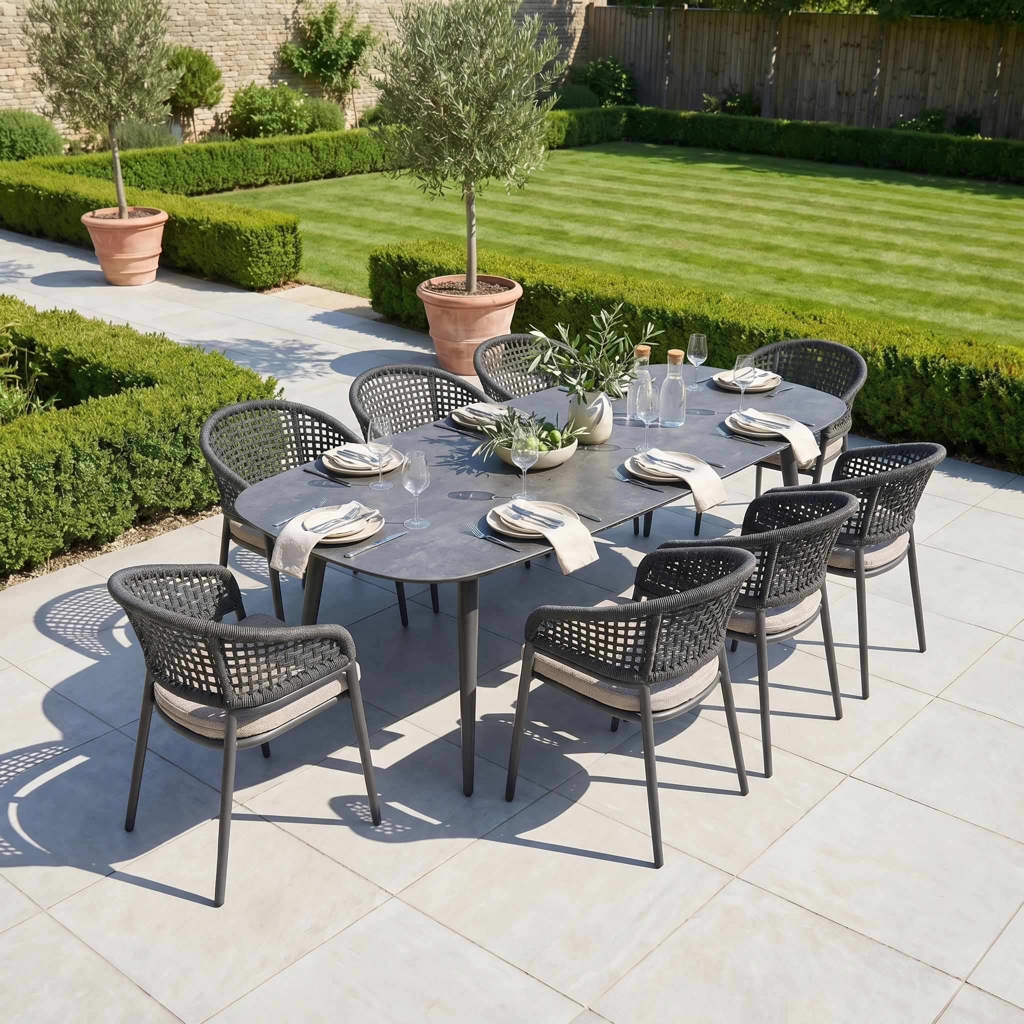 Outdoor dining set for eight on a patio with lush greenery. The Kalama 8 Seat Oval Rope Dining Set in Truffle offers stylish aluminium garden furniture, perfect for elegant gatherings outdoors.