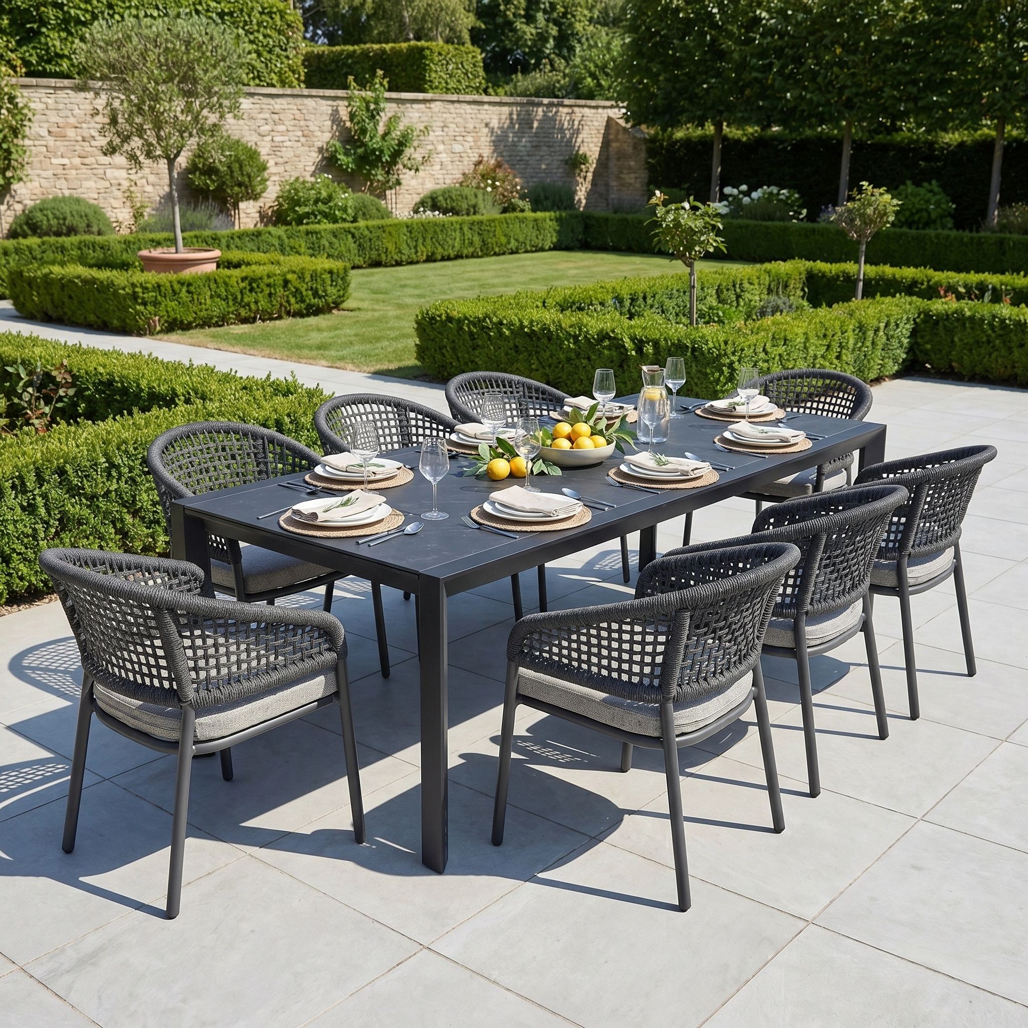 The Kalama 8 Seat Extending Dining Set with Ceramic Table in Truffle features sleek aluminium, set with plates, glasses, and fruit for stylish outdoor gatherings among lush greenery and hedges.