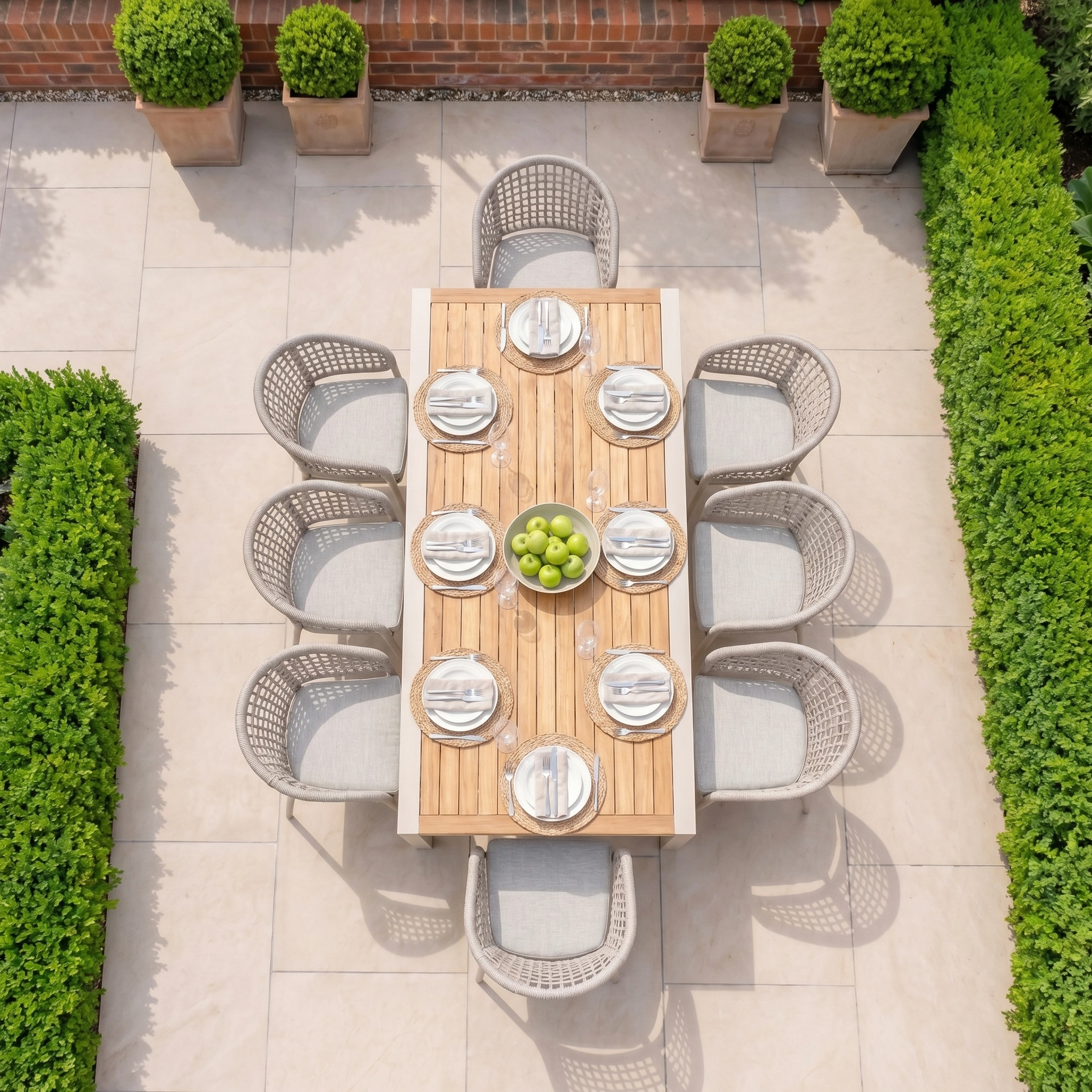 The Kalama 8 Seat Extending Dining Set with Teak Table in Oatmeal, topped with a bowl of green apples, is surrounded by lush greenery—ideal for stylish outdoor dining.