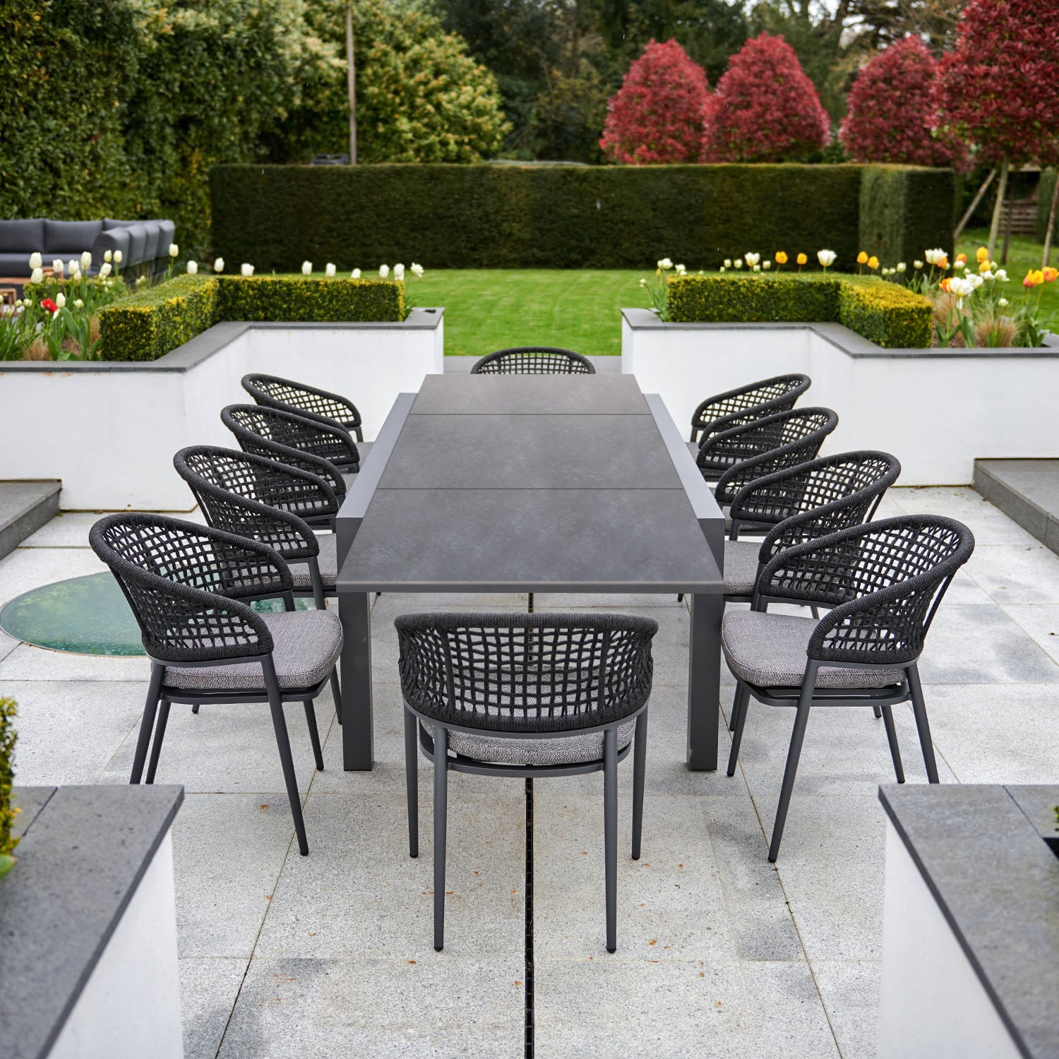 Modern outdoor dining area featuring the Kalama 8 Seat Rectangular Extending Dining Set with a ceramic table in charcoal and eight black woven chairs, all crafted from weather-proof aluminum, surrounded by a landscaped garden.