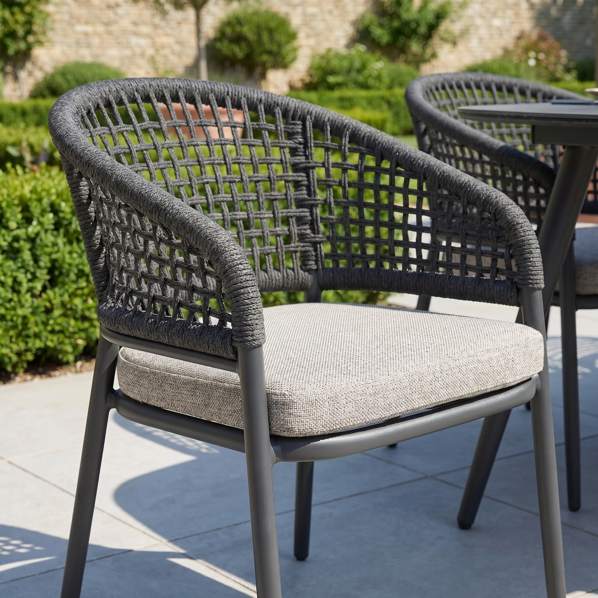 The Kalama 6 Seat Oval Rope Dining Set in Truffle sits on a patio, its modern woven design and light cushions complementing the garden furniture and lush greenery in the background.