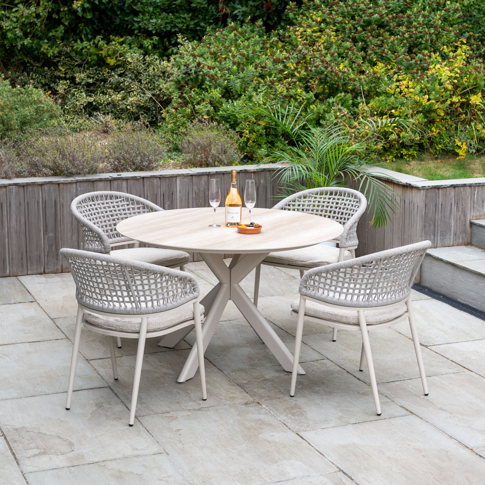 The Kalama 4 Seat Round Rope Dining Set with Albatross Table in Oatmeal offers stylish outdoor dining for four, perfect for relaxing meals on a stone terrace surrounded by greenery.