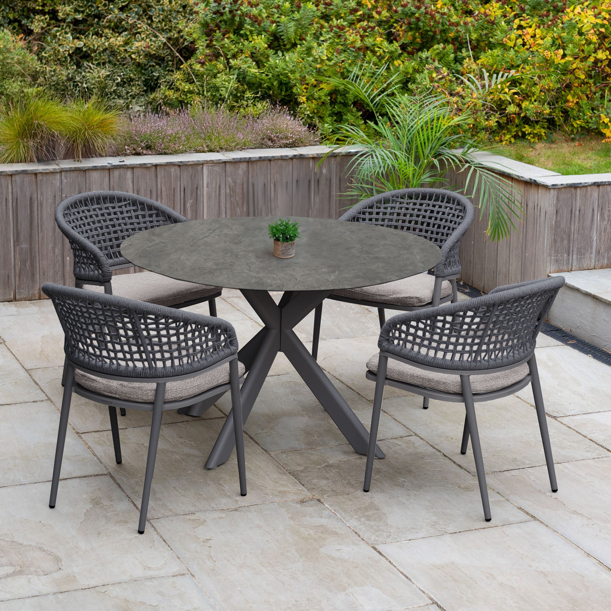 The Kalama 4 Seat Round Rope Dining Set with Albatross Table in Truffle includes a round outdoor table and four woven chairs—ideal for patios amid lush greenery.