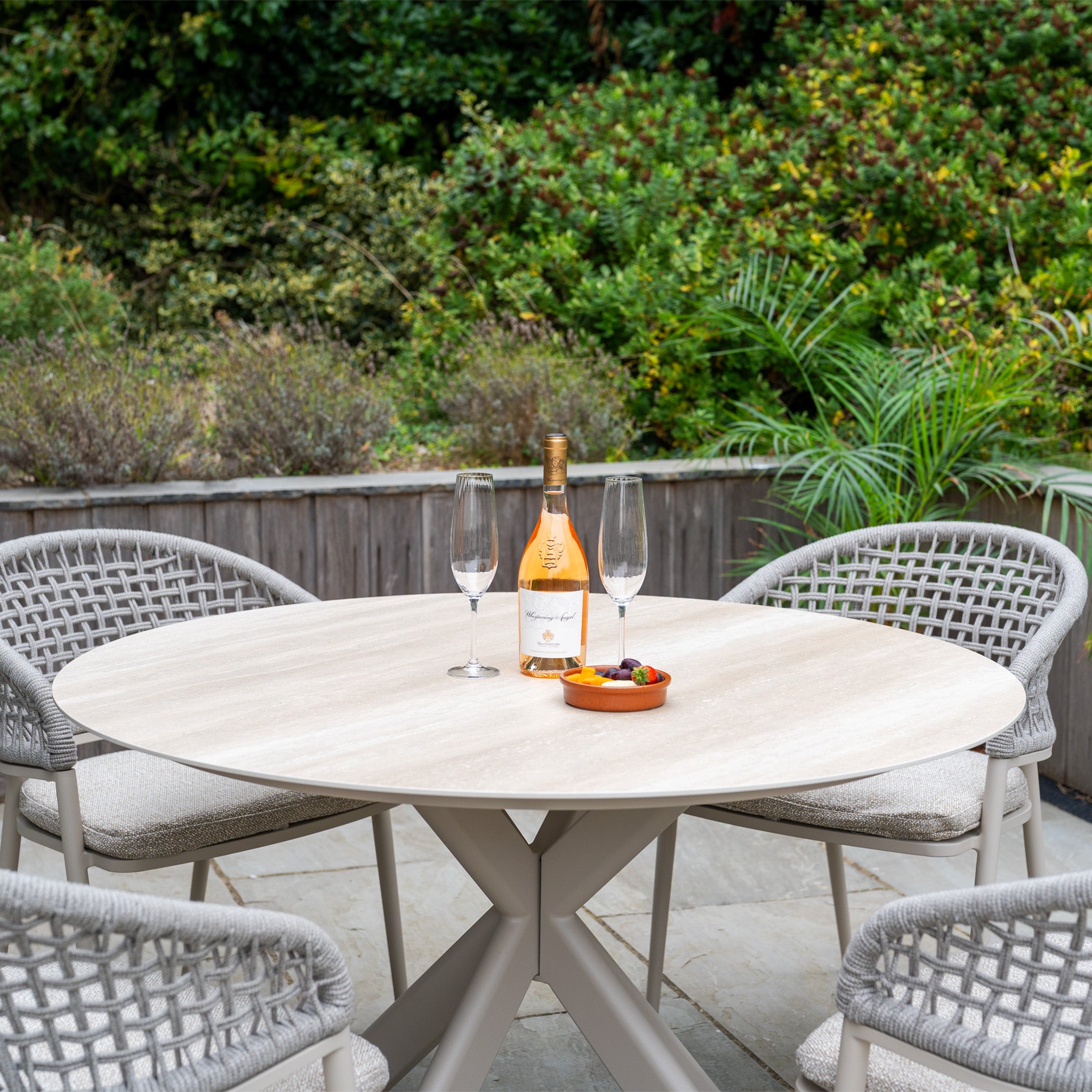 The Kalama 4 Seat Round Rope Dining Set with Albatross Table in Oatmeal creates a charming garden setup, featuring four chairs and a round table, perfect for enjoying drinks and snacks outdoors.