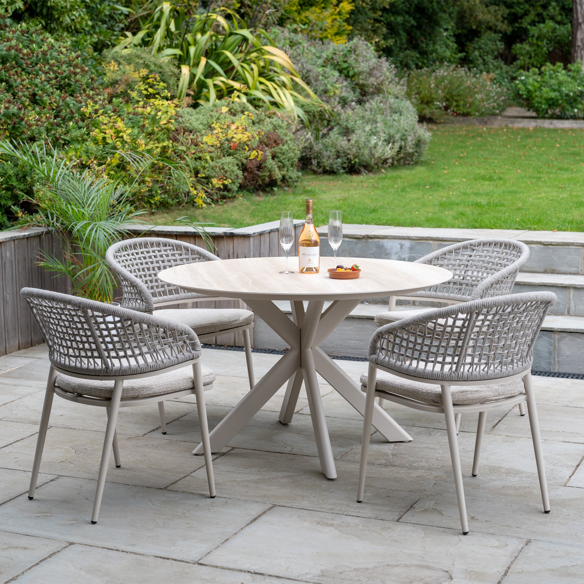 The Kalama 4 Seat Round Rope Dining Set with Albatross Table in Oatmeal is shown with four chairs, wine, glasses, and snacks arranged on the round patio table in a beautiful garden setting.