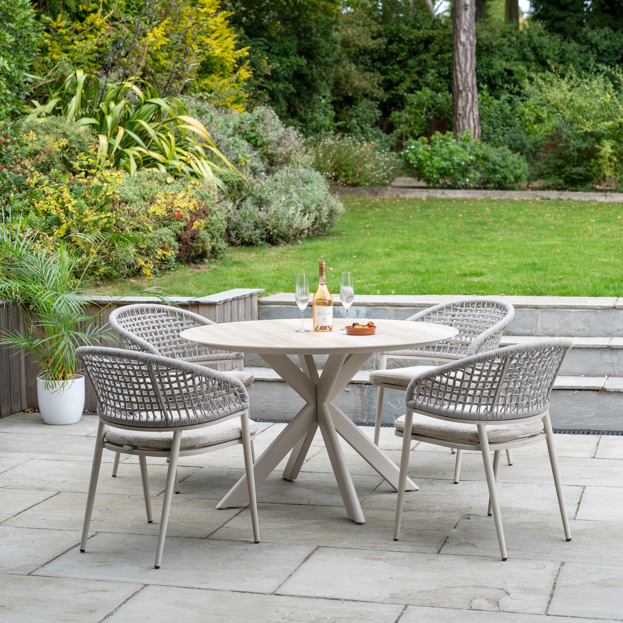 The Kalama 4 Seat Round Rope Dining Set with Albatross Table in Oatmeal creates an inviting garden dining spot on a stone patio, perfect for relaxing with wine and glasses in a lush outdoor setting.