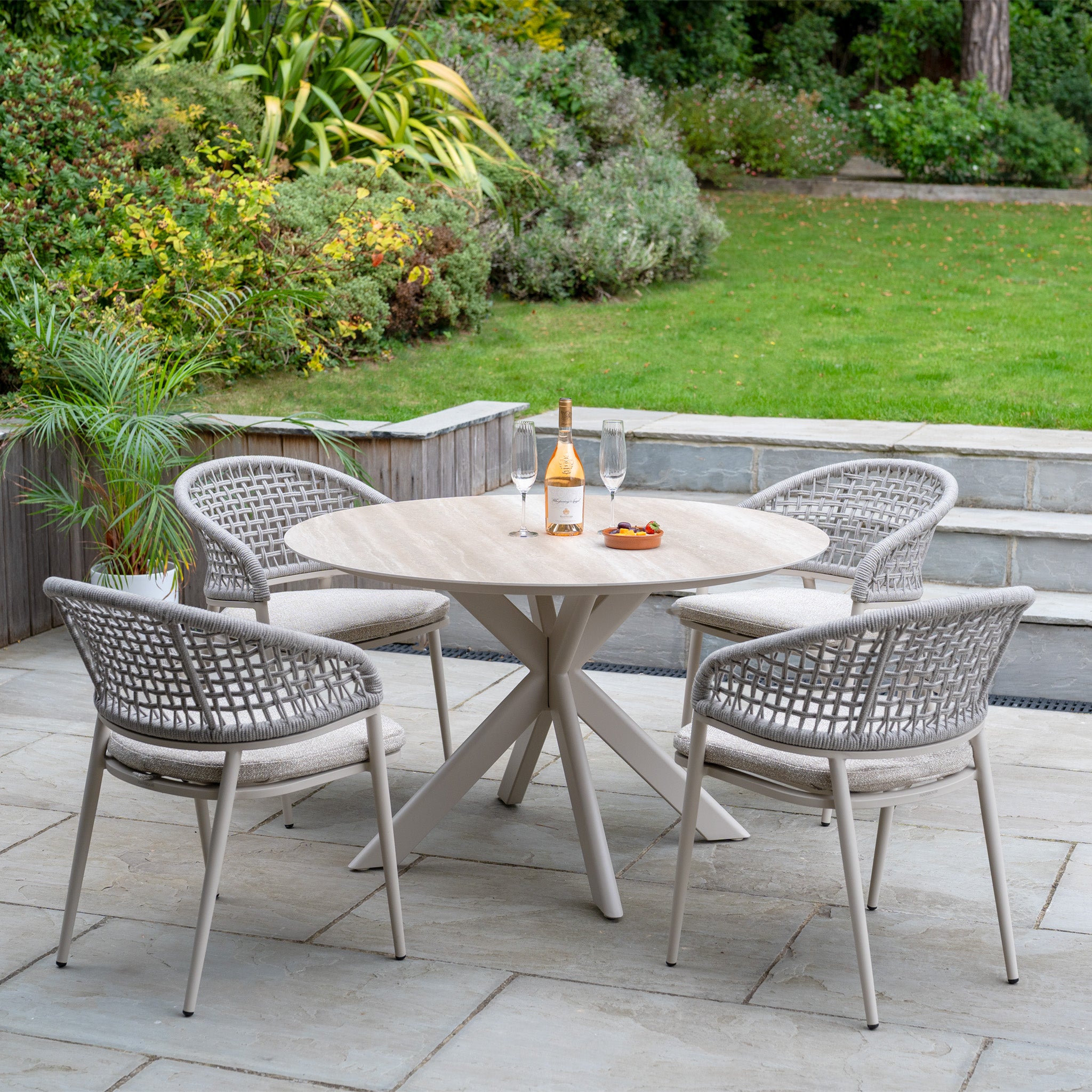 The Kalama 4 Seat Round Rope Dining Set with Albatross Table in Oatmeal is arranged with wine, glasses, and snacks for a stylish and relaxing garden dining experience outdoors.