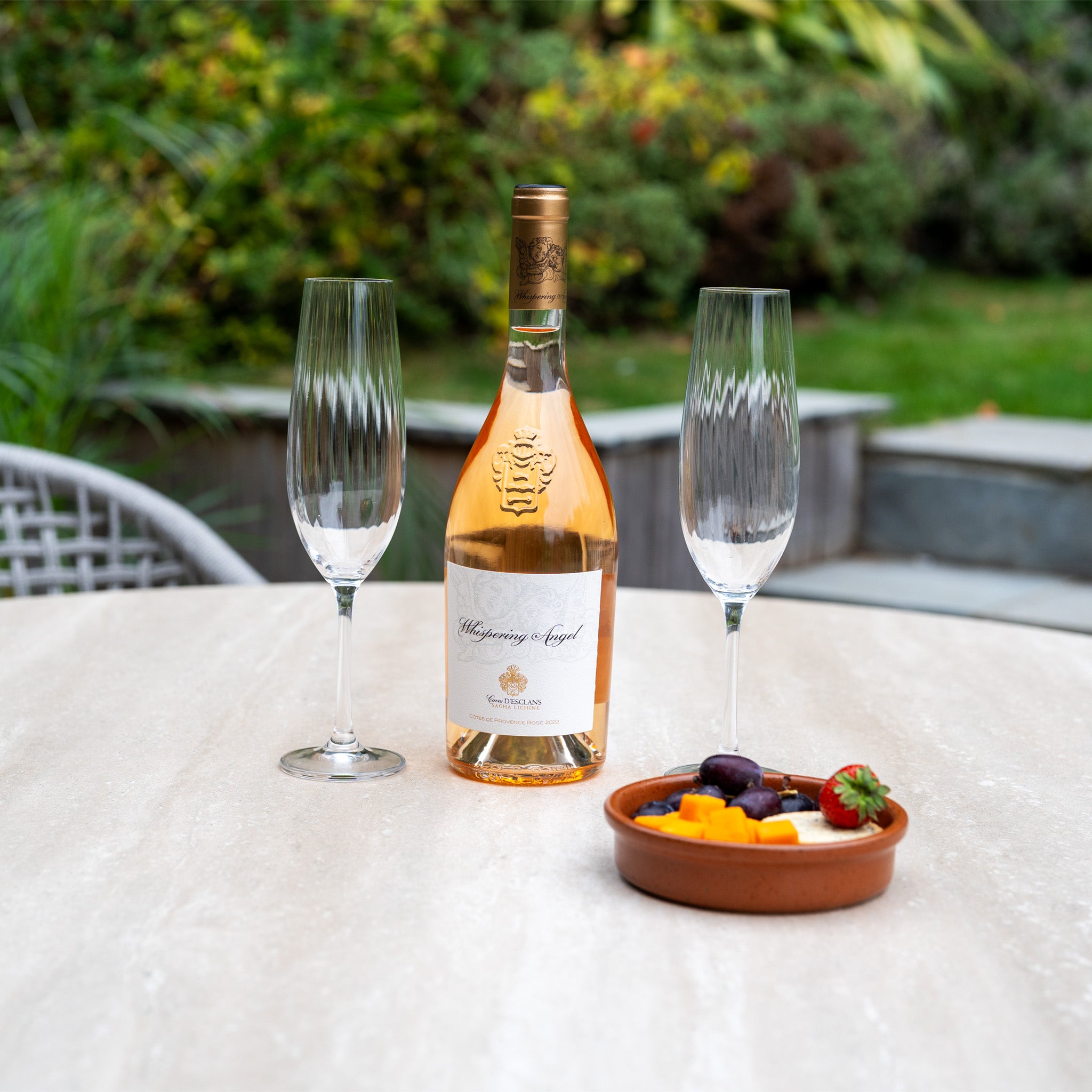 A bottle of rosé, two glasses, and a dish of fruit and cheese sit atop the Kalama 4 Seat Round Rope Dining Set with Albatross Table in Oatmeal on an outdoor patio.