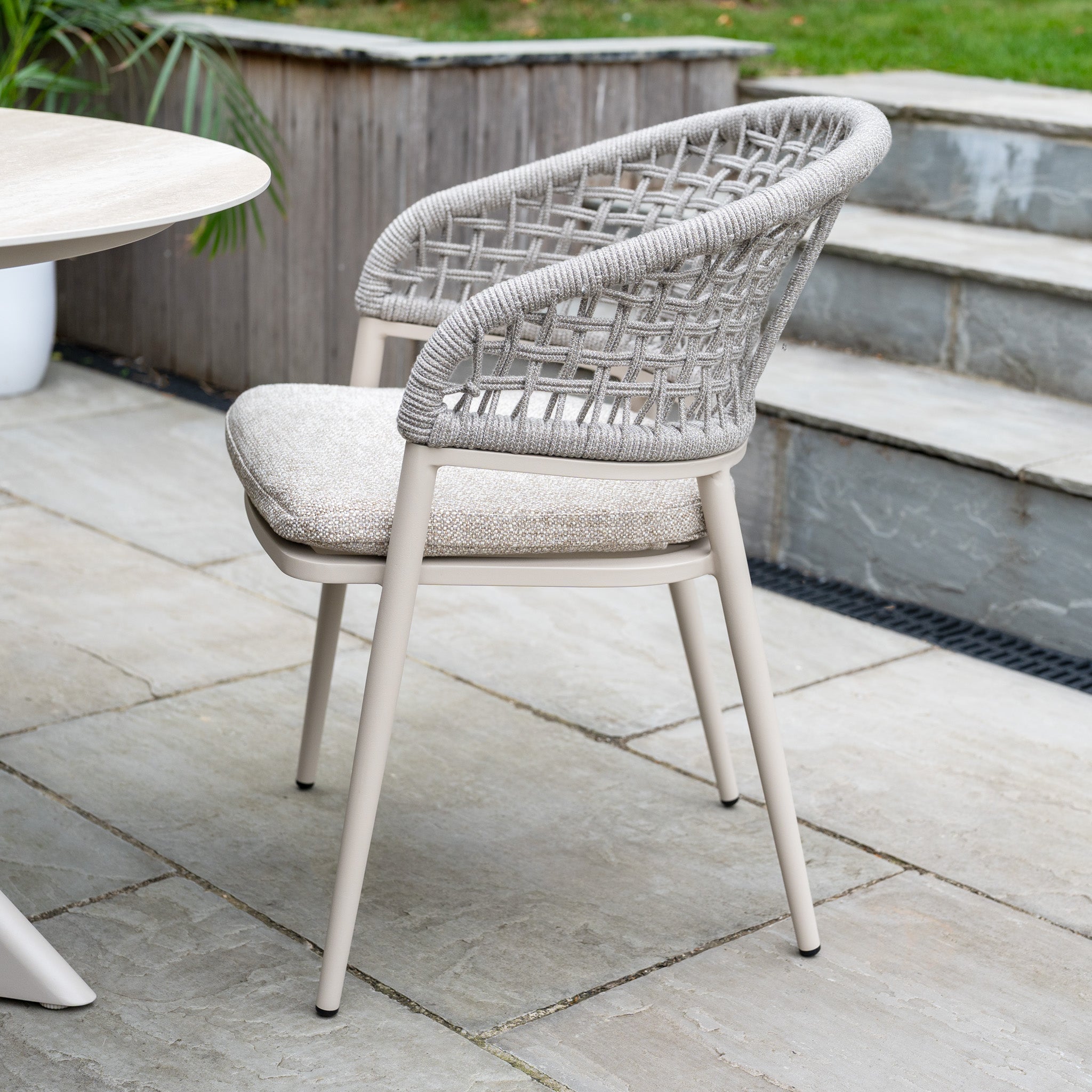 The Kalama 4 Seat Round Rope Dining Set with Albatross Table in Oatmeal features woven-back patio chairs and cushioned seats, creating a stylish garden dining set ideal for stone patios surrounded by greenery.