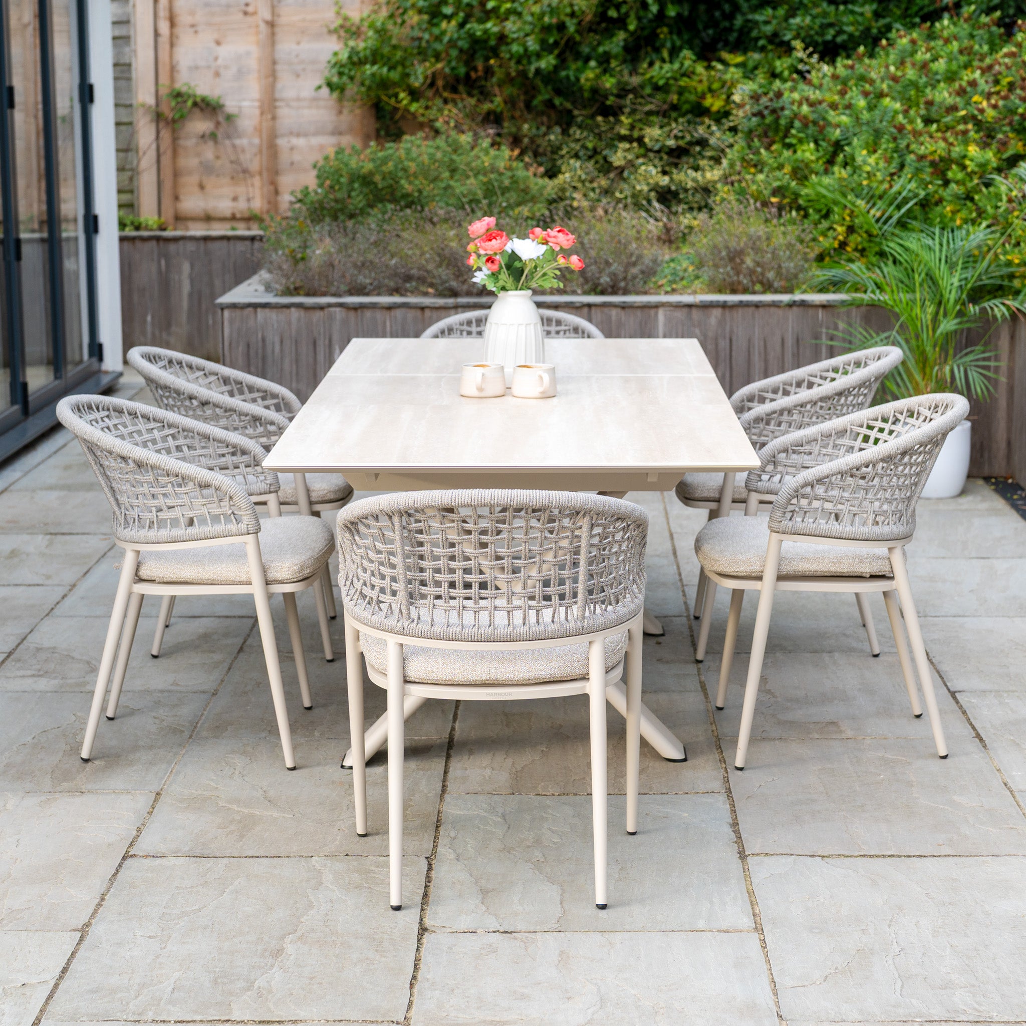 The Kalama 6 Seat Rope Dining Set with Albatross Extending Table in Oatmeal features a stylish outdoor table and six rope-woven chairs, beautifully arranged on a stone patio surrounded by greenery.