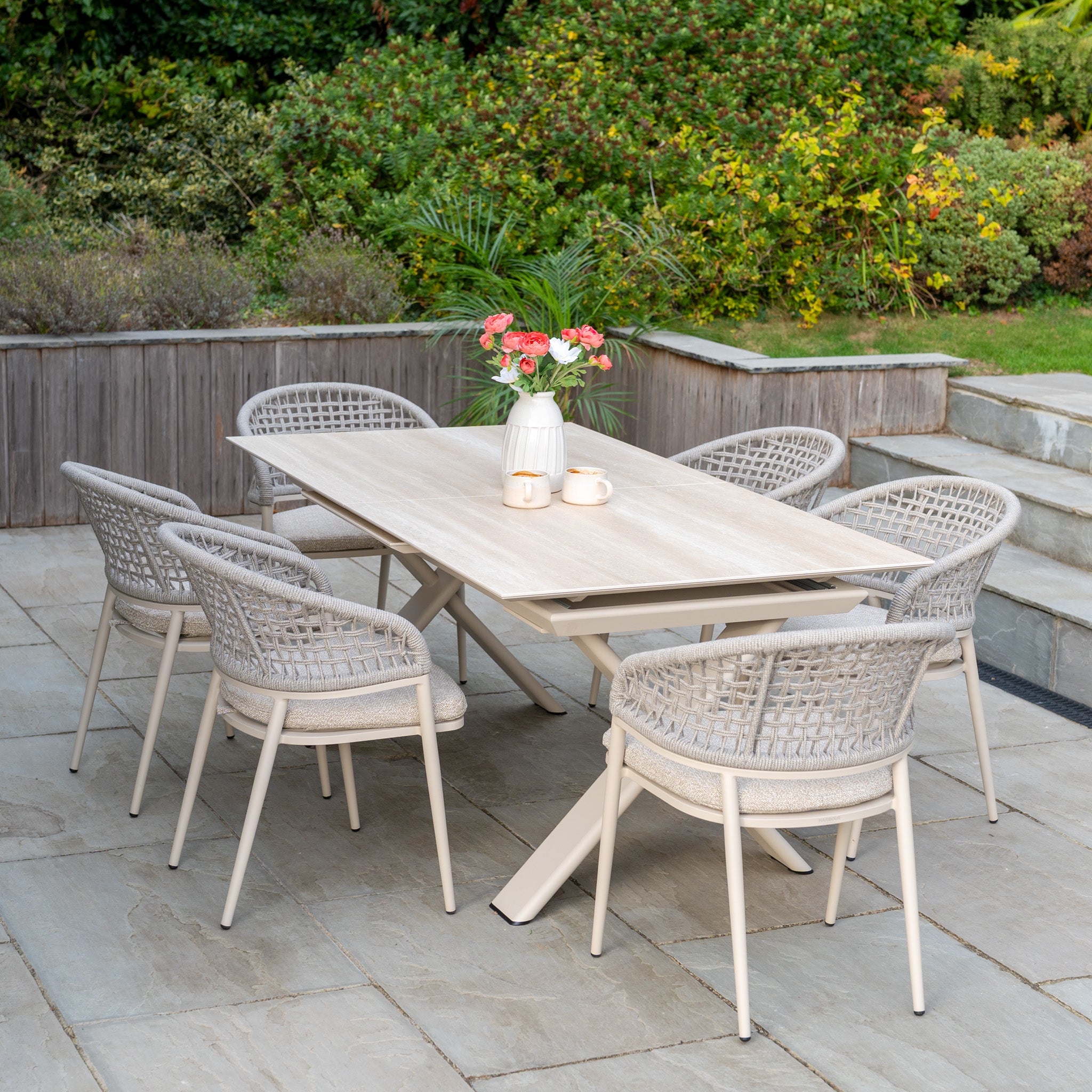 Outdoor patio features the Kalama 6 Seat Rope Dining Set with Albatross Extending Table in Oatmeal, six chairs, a white vase of flowers, and two cups on a stone terrace.