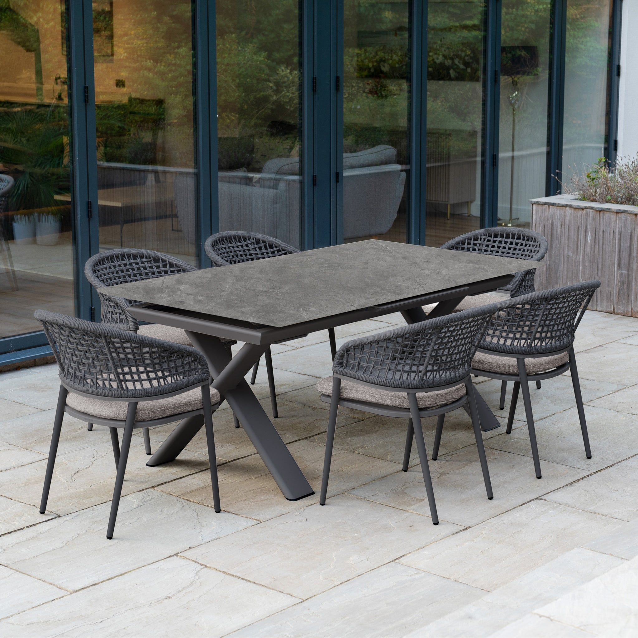 The Kalama 6 Seat Rope Dining Set with Albatross Extending Table in Truffle offers stylish outdoor dining, featuring six woven chairs on a stone patio by glass doors—perfect for modern gatherings.