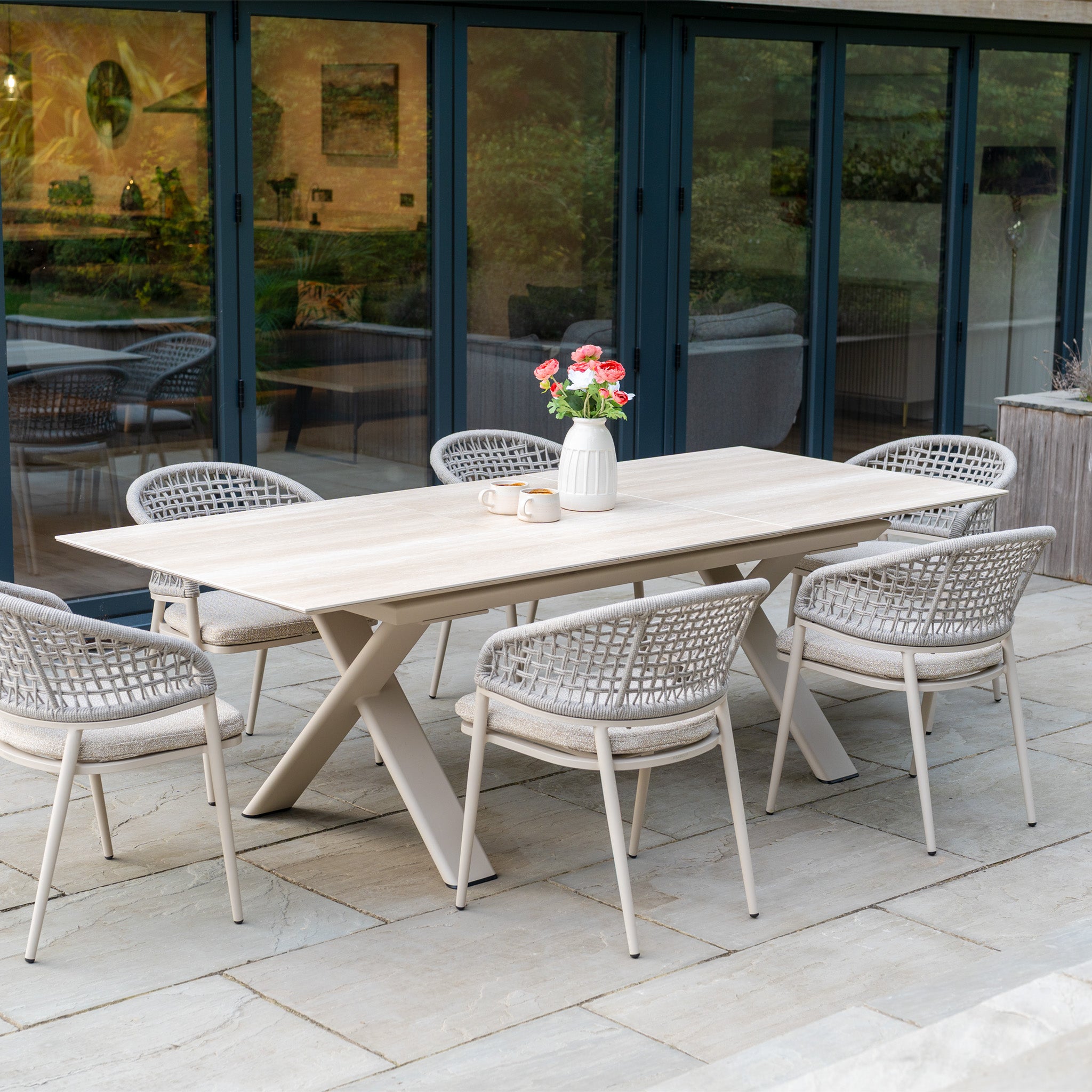 The Kalama 6 Seat Rope Dining Set with Albatross Extending Table in Oatmeal features six woven chairs on a stone patio, accented with outdoor boucle fabric and a vase of flowers for a modern touch.