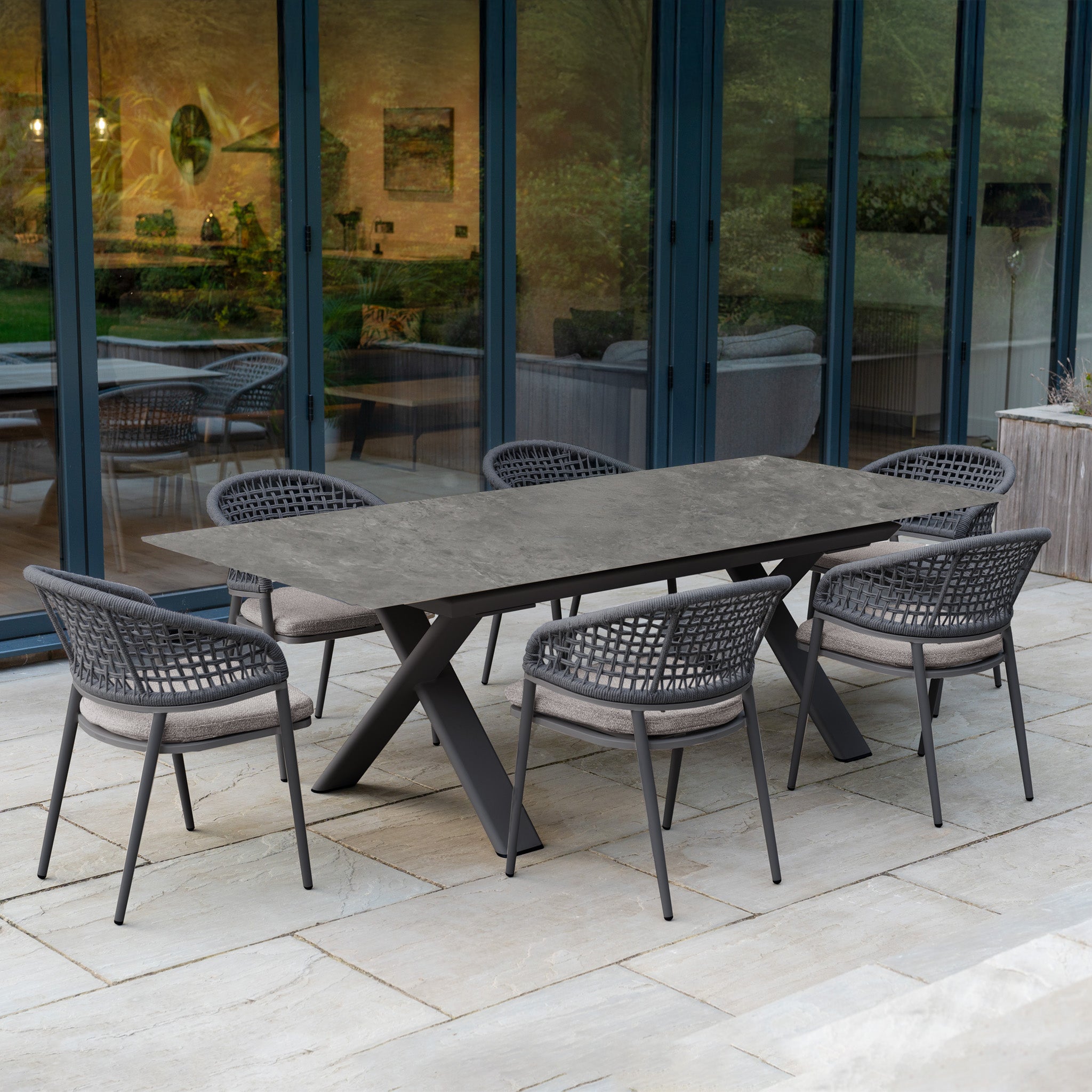Modern outdoor dining with the Kalama 6 Seat Rope Dining Set featuring an Albatross Extending Table in Truffle, showcased on a stone patio near glass doors.