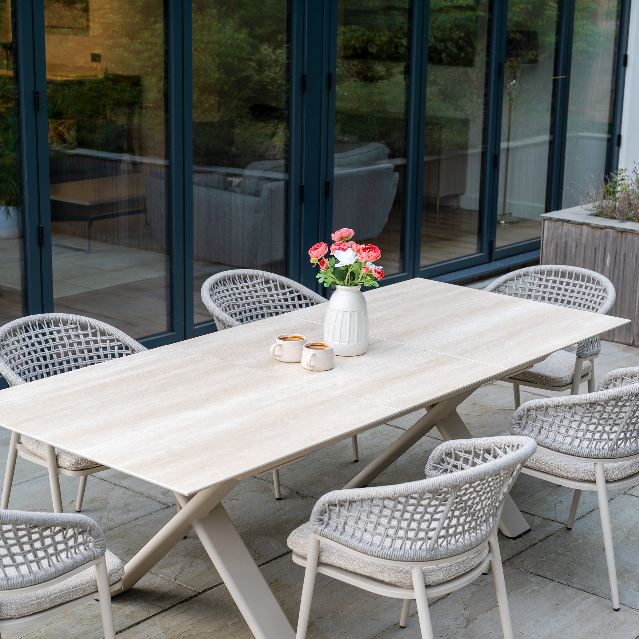 The Kalama 6 Seat Rope Dining Set with Albatross Extending Table in Oatmeal features six chairs with outdoor boucle fabric and is styled with two mugs and a vase of pink flowers on top.