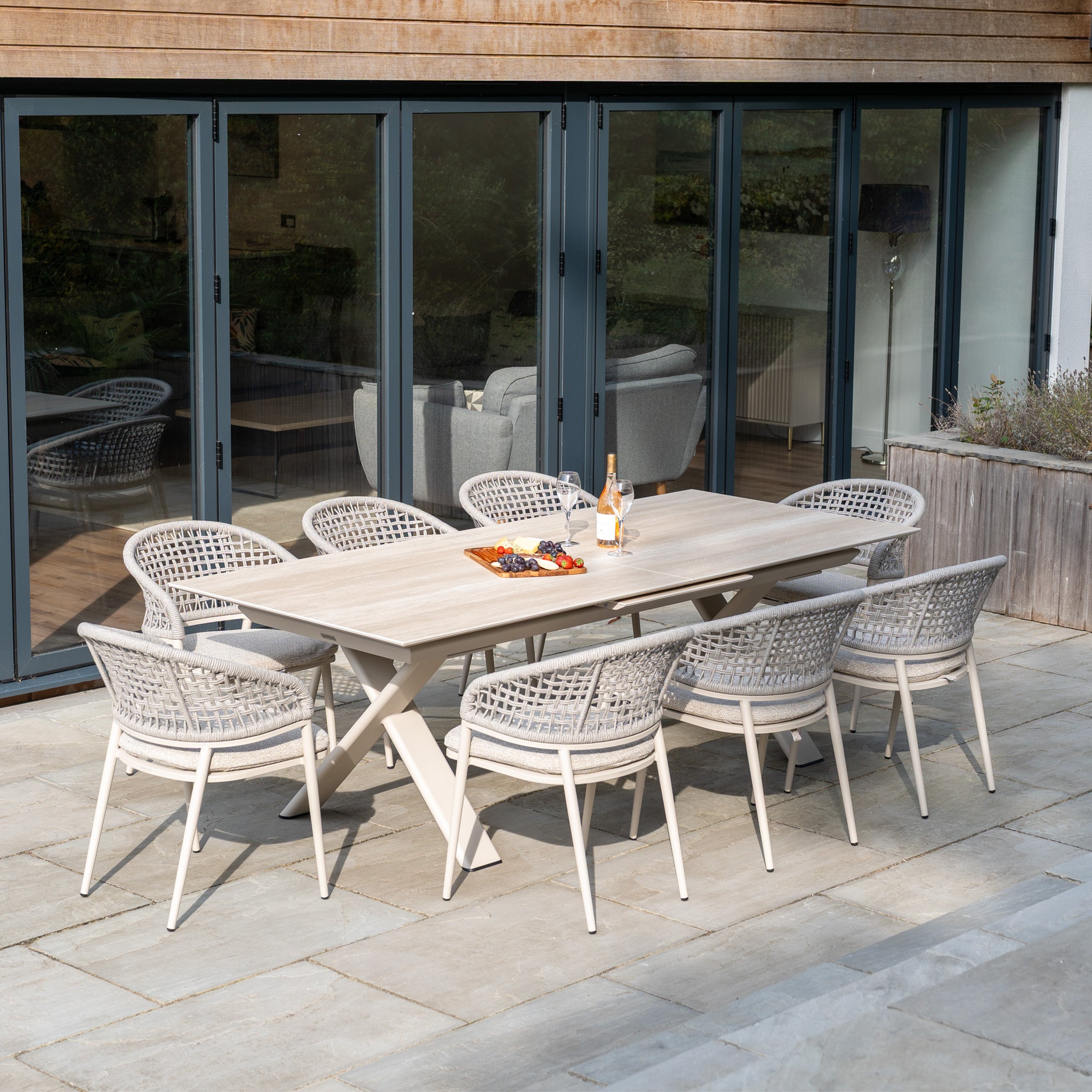 The Kalama 8 Seat Rope Dining Set with Albatross Extending Table in Oatmeal is arranged with drinks and snacks on a stone patio near glass doors for stylish outdoor dining.