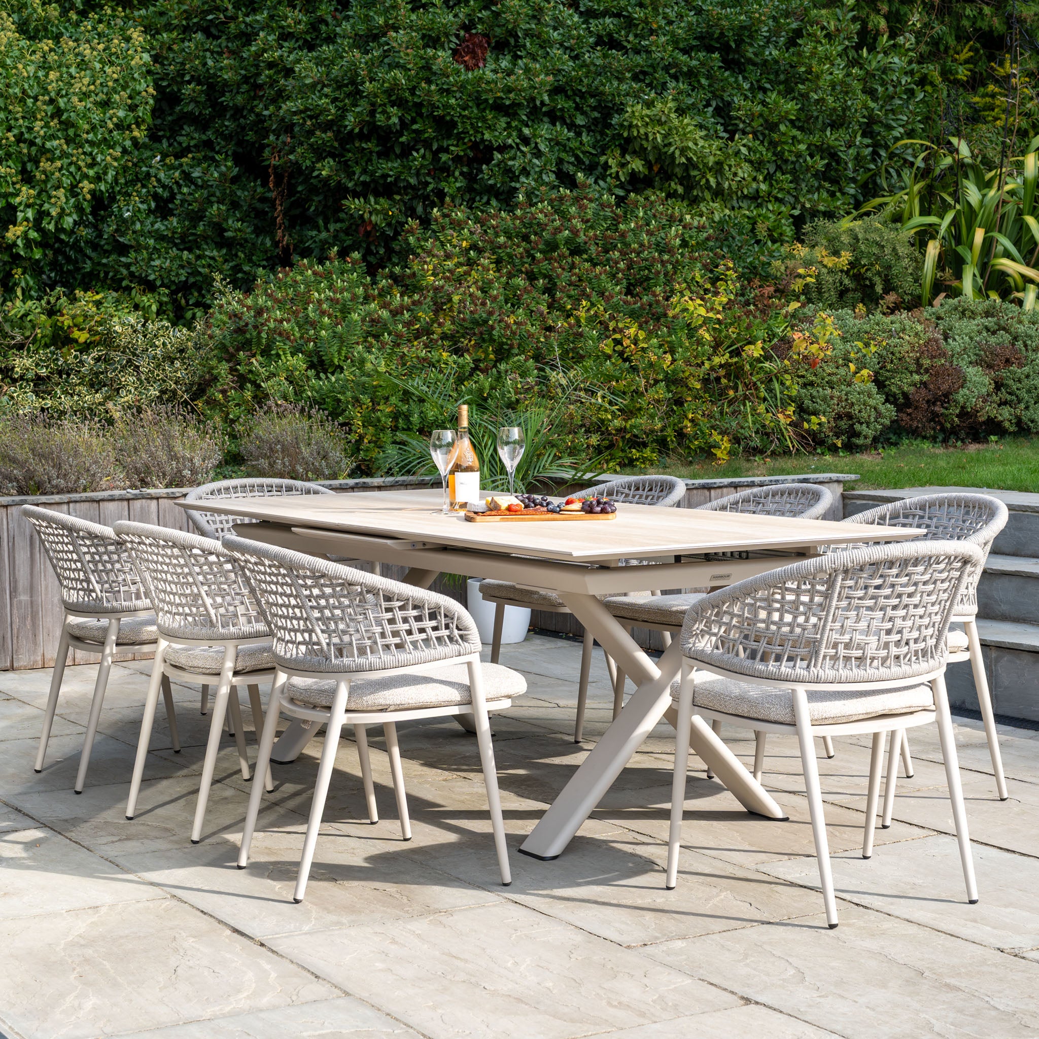 Enjoy alfresco meals with the Kalama 8 Seat Rope Dining Set featuring an Albatross Extending Table in Oatmeal—perfect for gatherings on a stone patio surrounded by lush greenery.