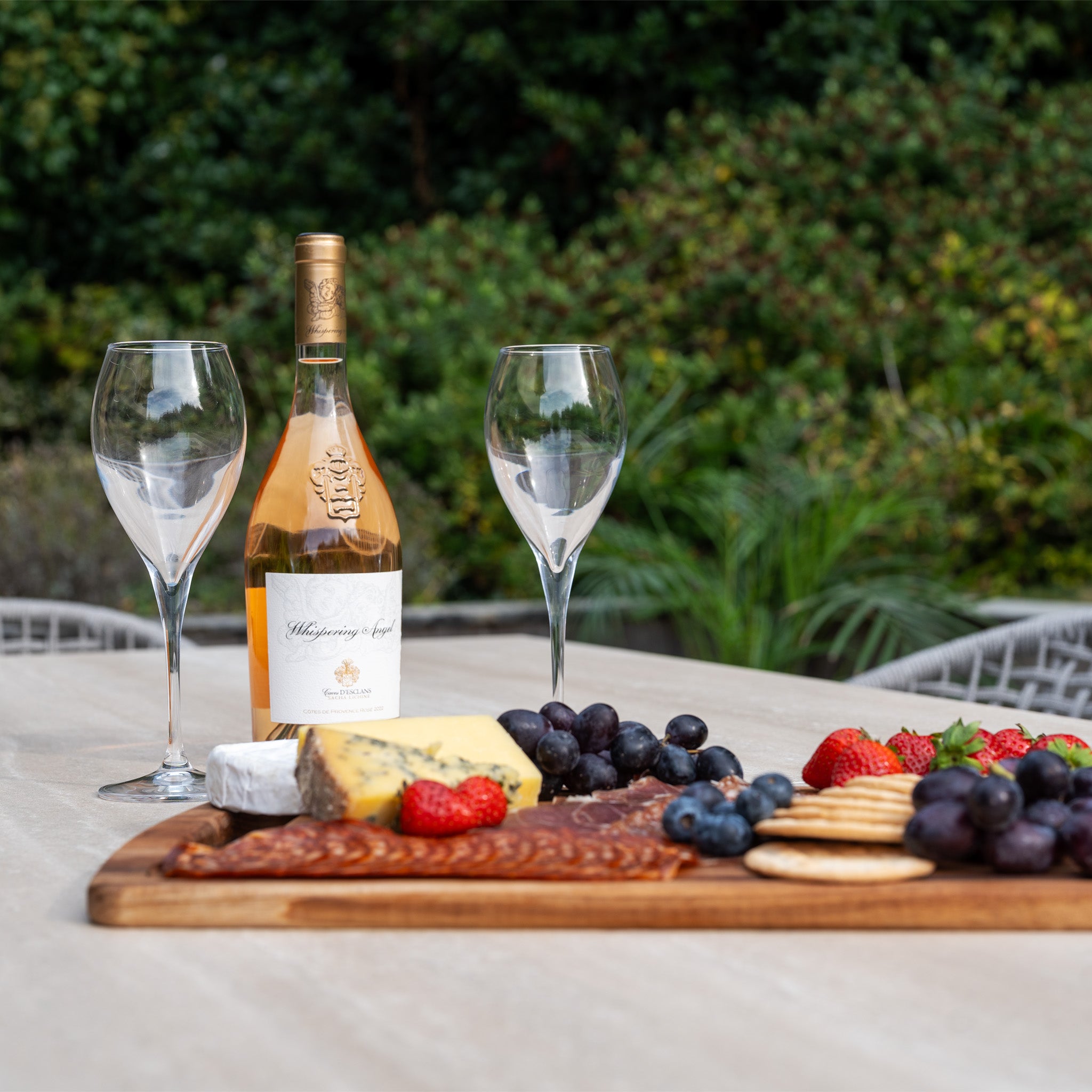A bottle of rosé, two wine glasses, and a cheese and fruit platter sit atop the Kalama 6 Seat Rope Dining Set with Albatross Extending Table in Oatmeal, creating an inviting space to relax in style.