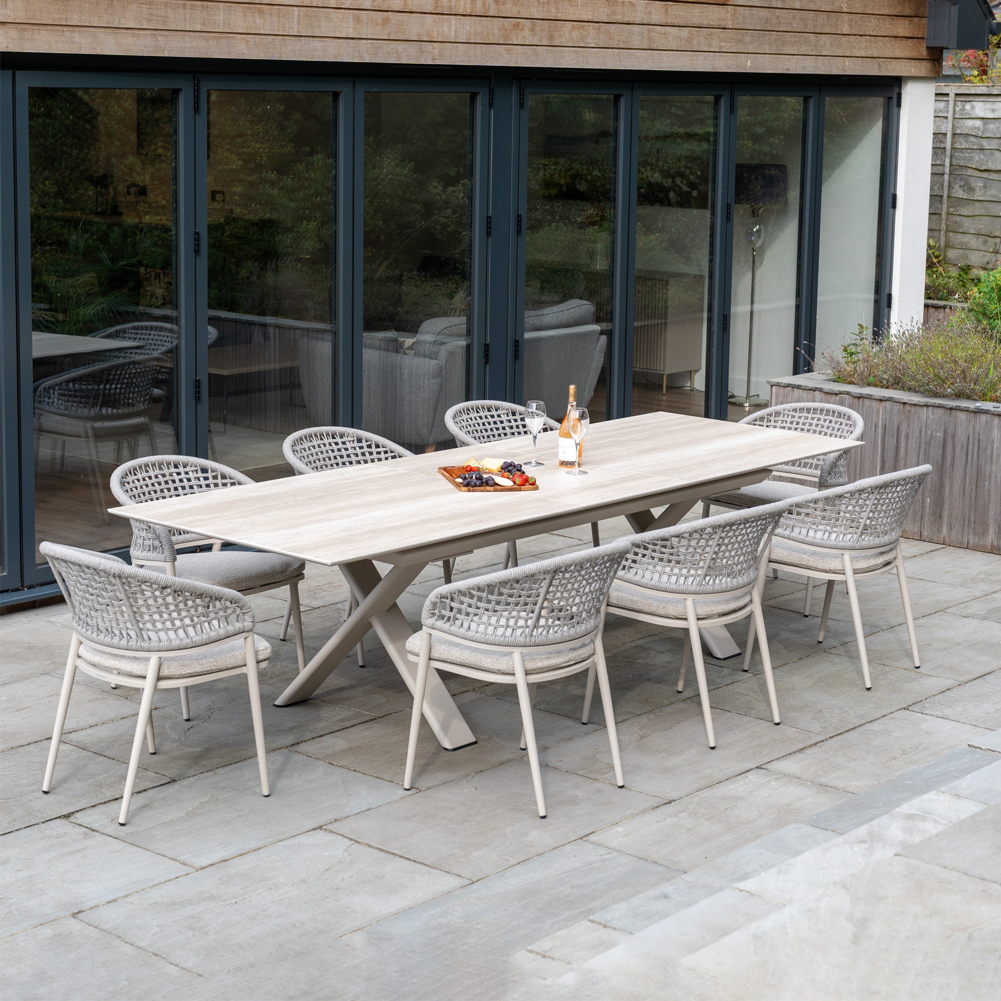 The Kalama 8 Seat Rope Dining Set with Albatross Extending Table in Oatmeal showcases an outdoor table and eight woven chairs, arranged with drinks and snacks on a stone patio by glass doors.