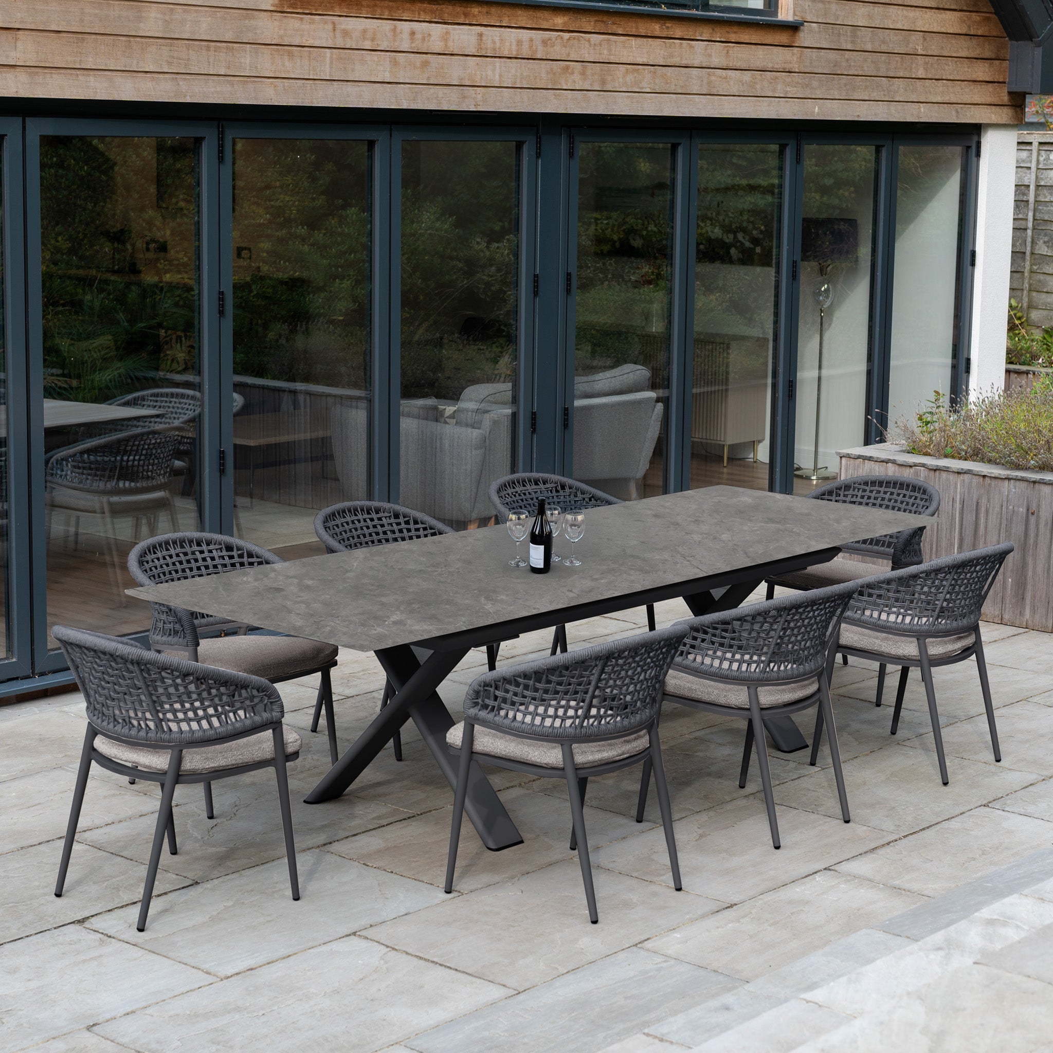 The Kalama 8 Seat Rope Dining Set with Albatross Extending Table in Truffle features eight woven chairs around a stylish patio table, elegantly set with a wine bottle and glasses.