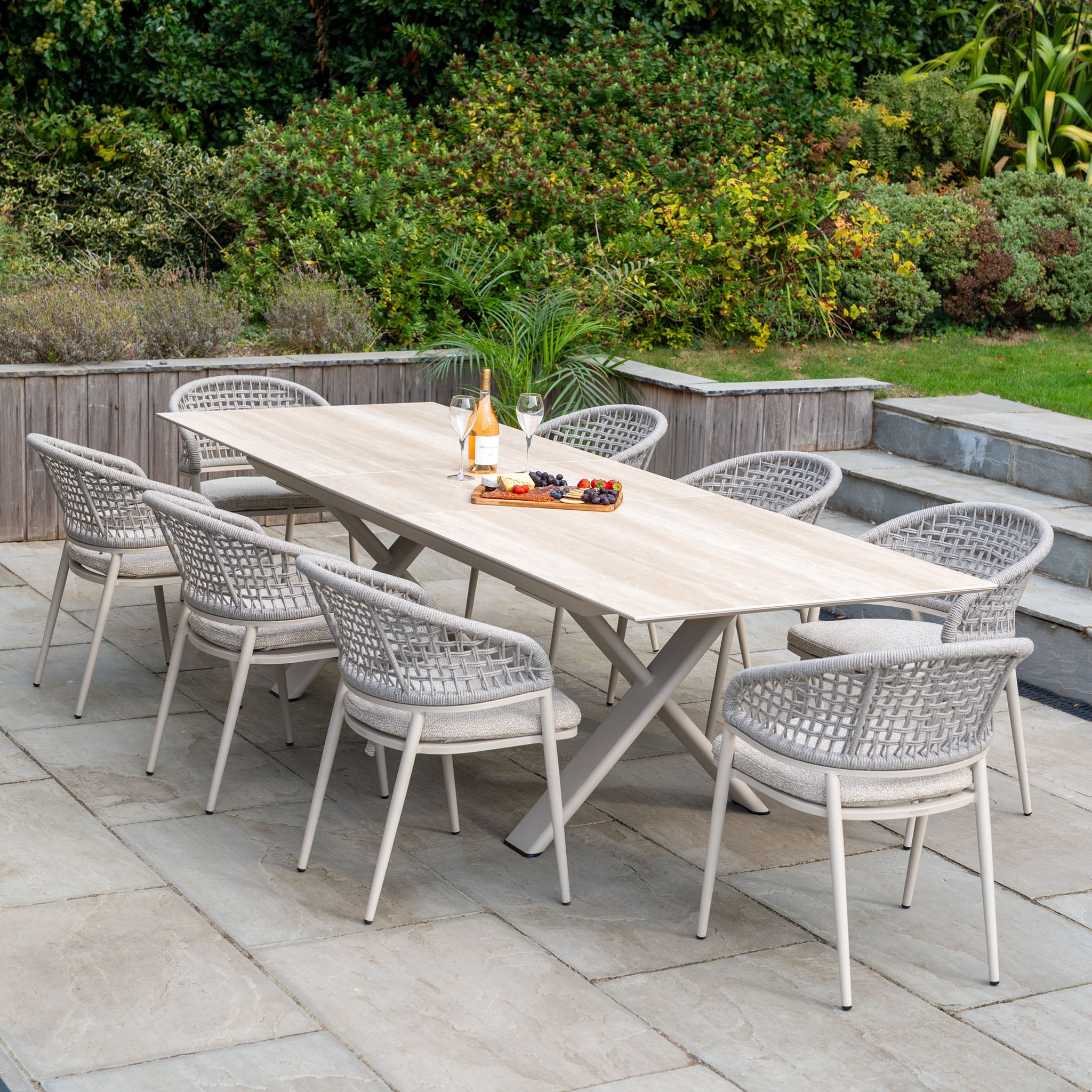 The Kalama 8 Seat Rope Dining Set with Albatross Extending Table in Oatmeal creates an elegant space for memorable outdoor gatherings, surrounded by greenery on a stone patio with wine and snacks.