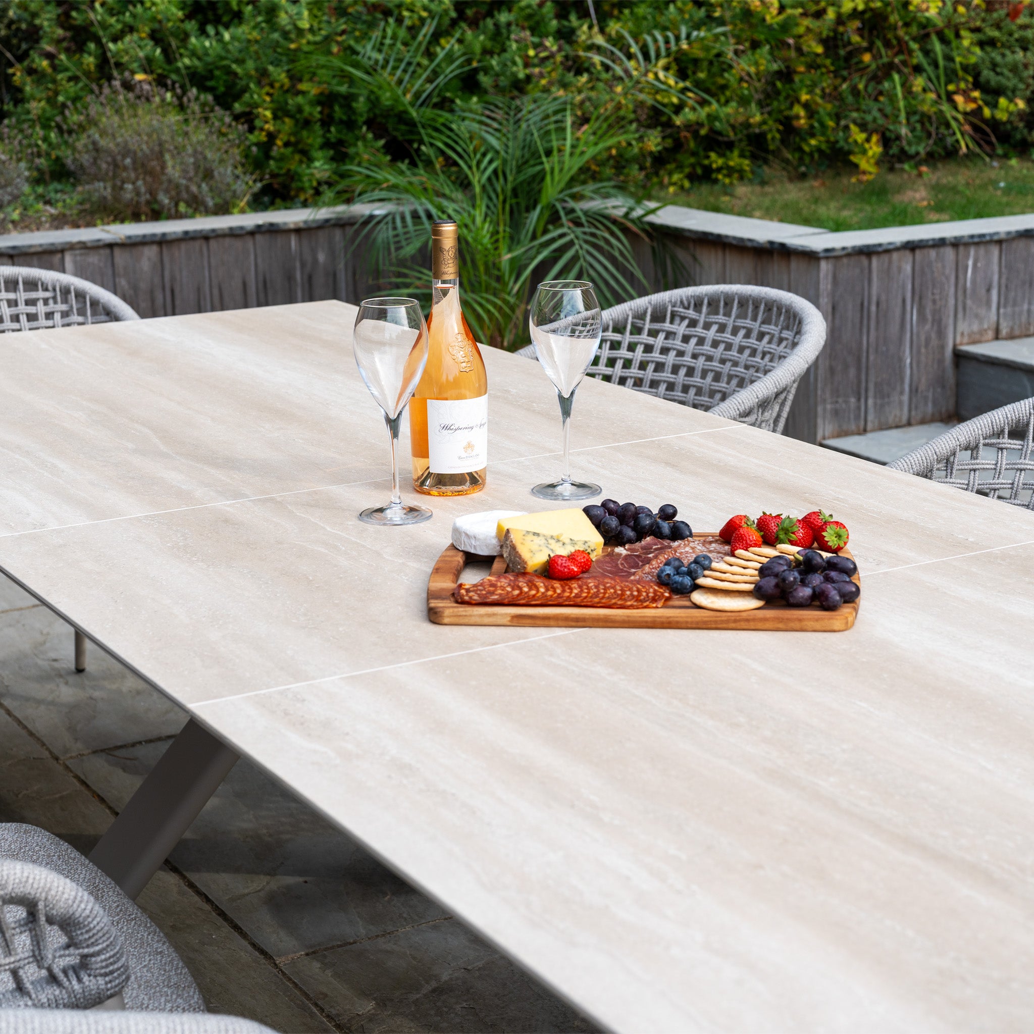 A table with wine, cheese, crackers, fruit, and salami on a wooden board outdoors—perfect for enjoying with the Kalama 8 Seat Rope Dining Set with Albatross Extending Table in Oatmeal or other stylish outdoor dining furniture.