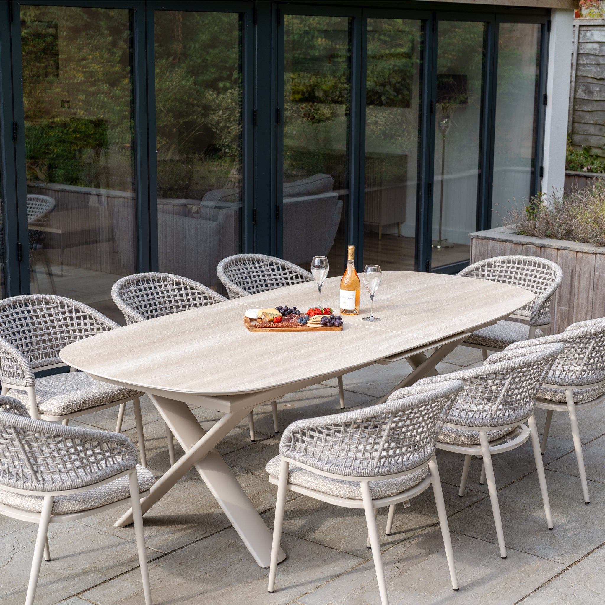 Outdoor dining table with eight chairs, wine, glasses, and a cheese platter on a stone patio by glass doors—the perfect setting for gatherings with the Kalama 8 Seat Oval Rope Dining Set with Albatross Extending Table in Oatmeal.